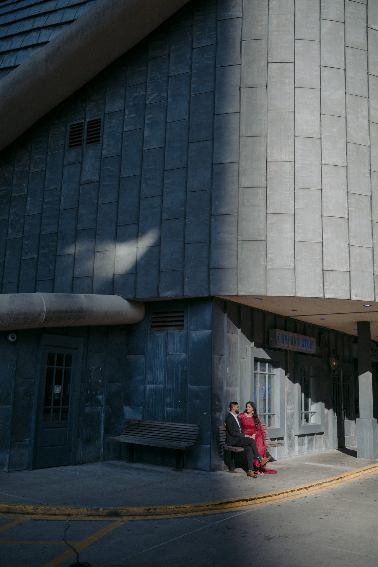 A couple in red and dark attire sits on a bench outside a large, dark paneled building.
