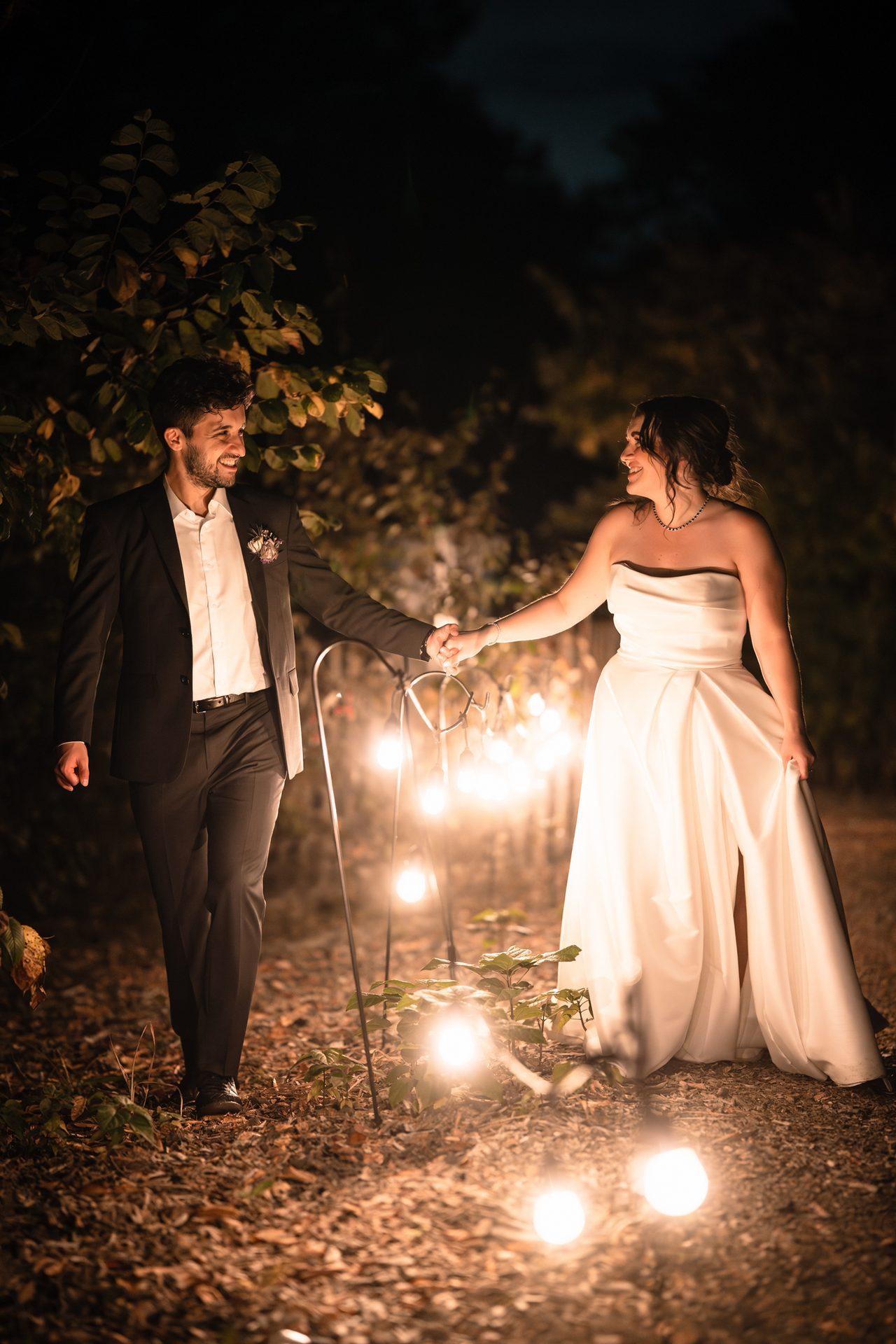 A joyful couple holding hands at night, illuminated by warm string lights, smiling at each other.