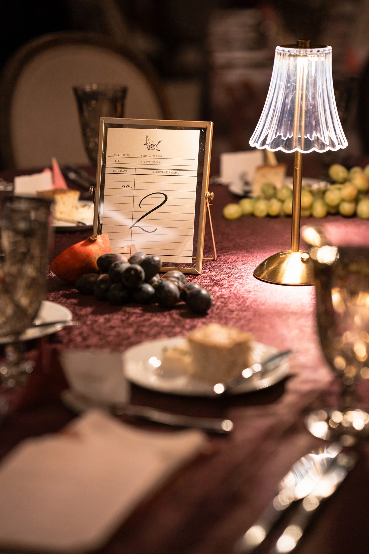Gold-framed table number '2' for 'Muna & Trevor', a small lamp, and fruit on a burgundy table.