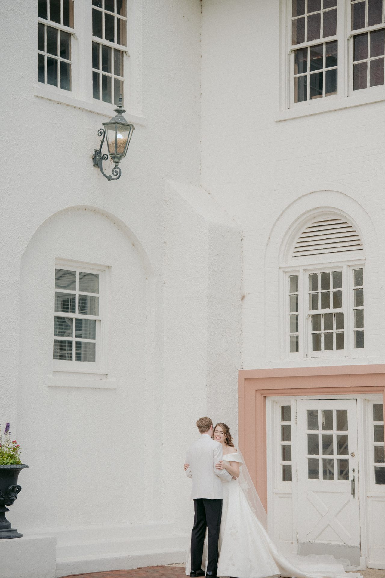 Bride and groom embracing outside a white building, the bride smiling at the camera.