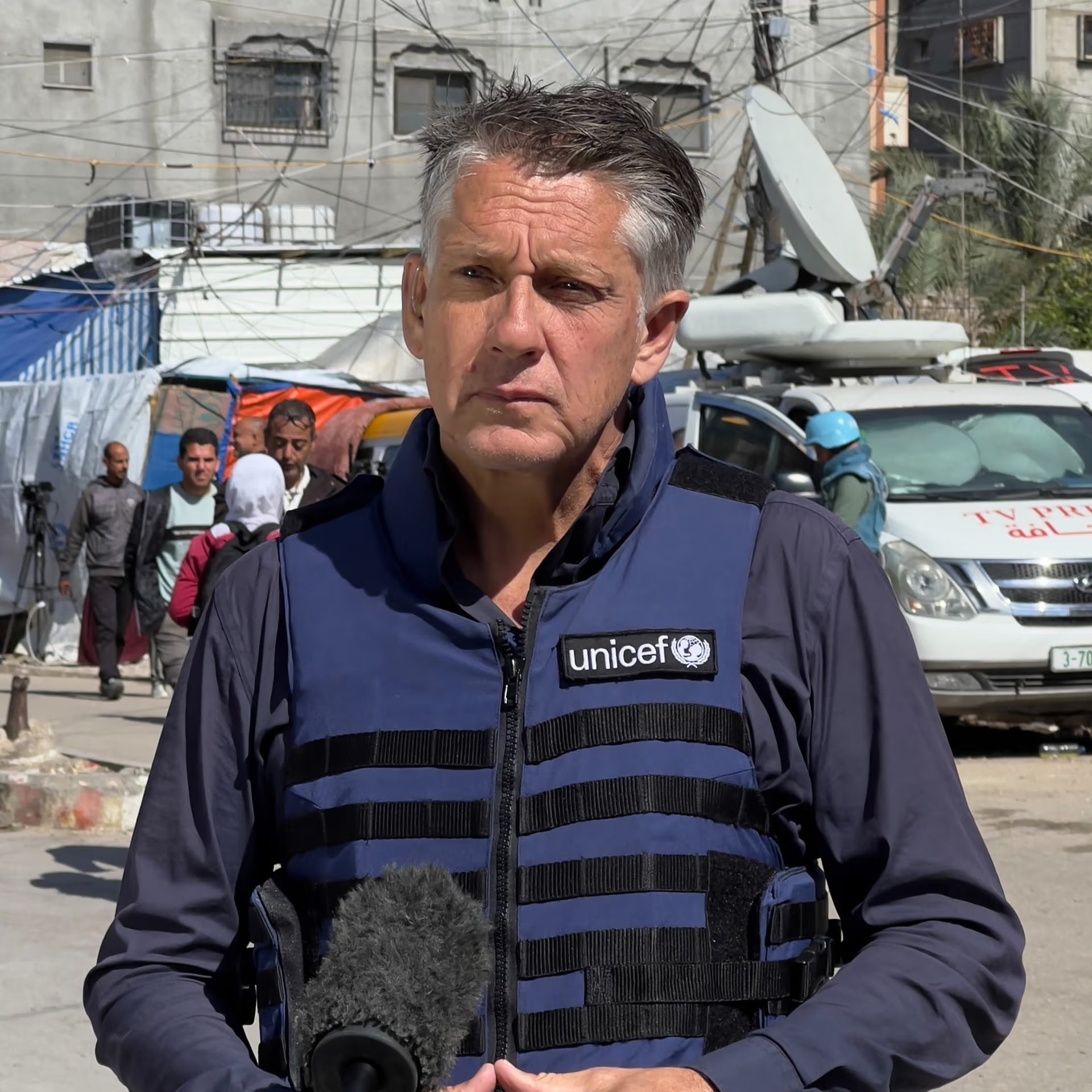 A man in a UNICEF bulletproof vest holds a microphone, looking serious, with people and a TV press vehicle in the background.