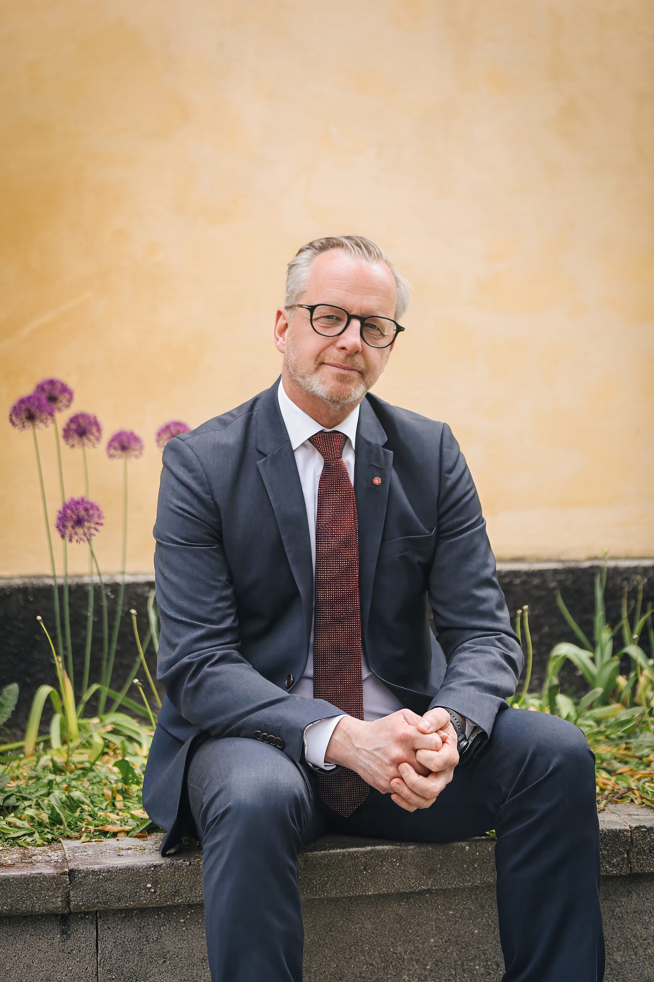A confident man in a dark suit, glasses, and red tie sits on a stone ledge against a yellow wall.