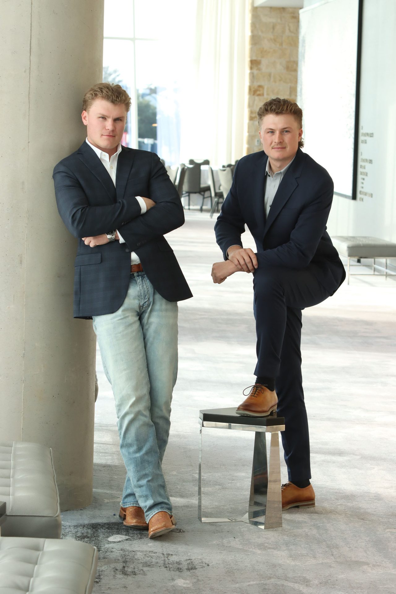 Two young men in blazers, one in jeans, one with foot on a stool, posing in a modern lobby.