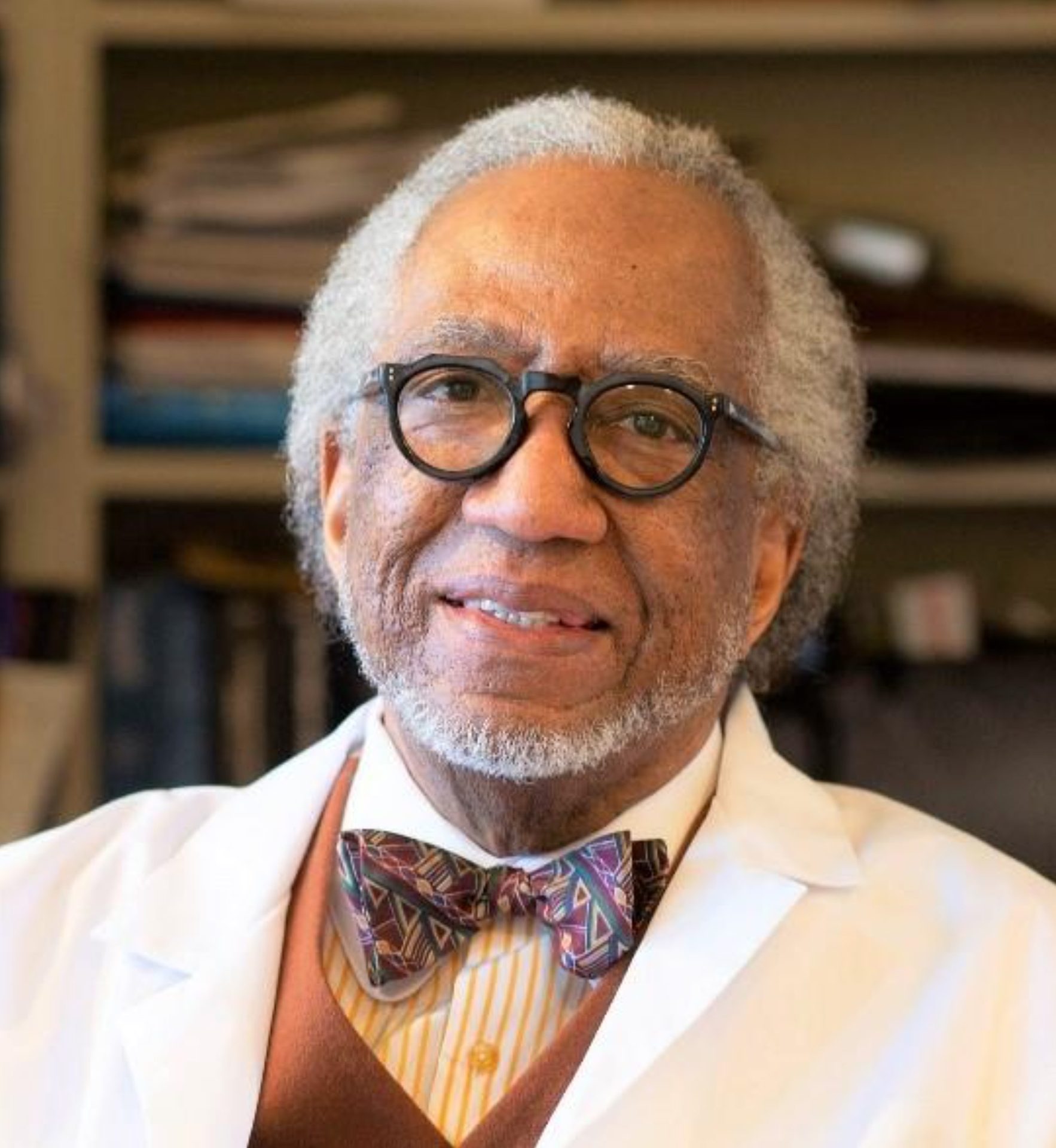 An older Black man with gray hair and glasses, wearing a lab coat and bow tie, smiles.