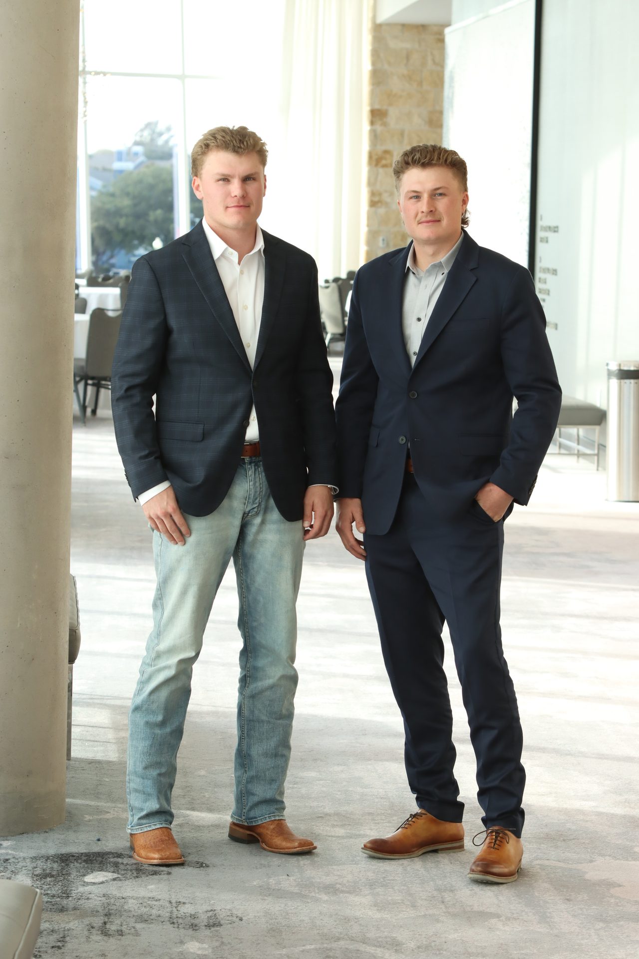 Two young men in suits, one in jeans, standing indoors and looking at the camera.