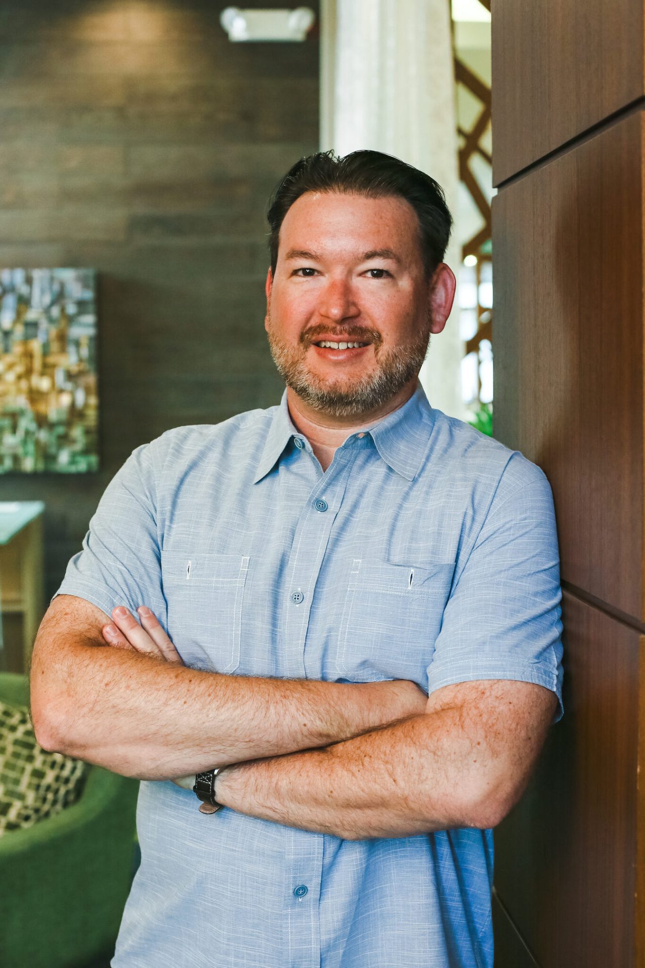 Man with beard and blue shirt, arms crossed, smiling in a modern interior.