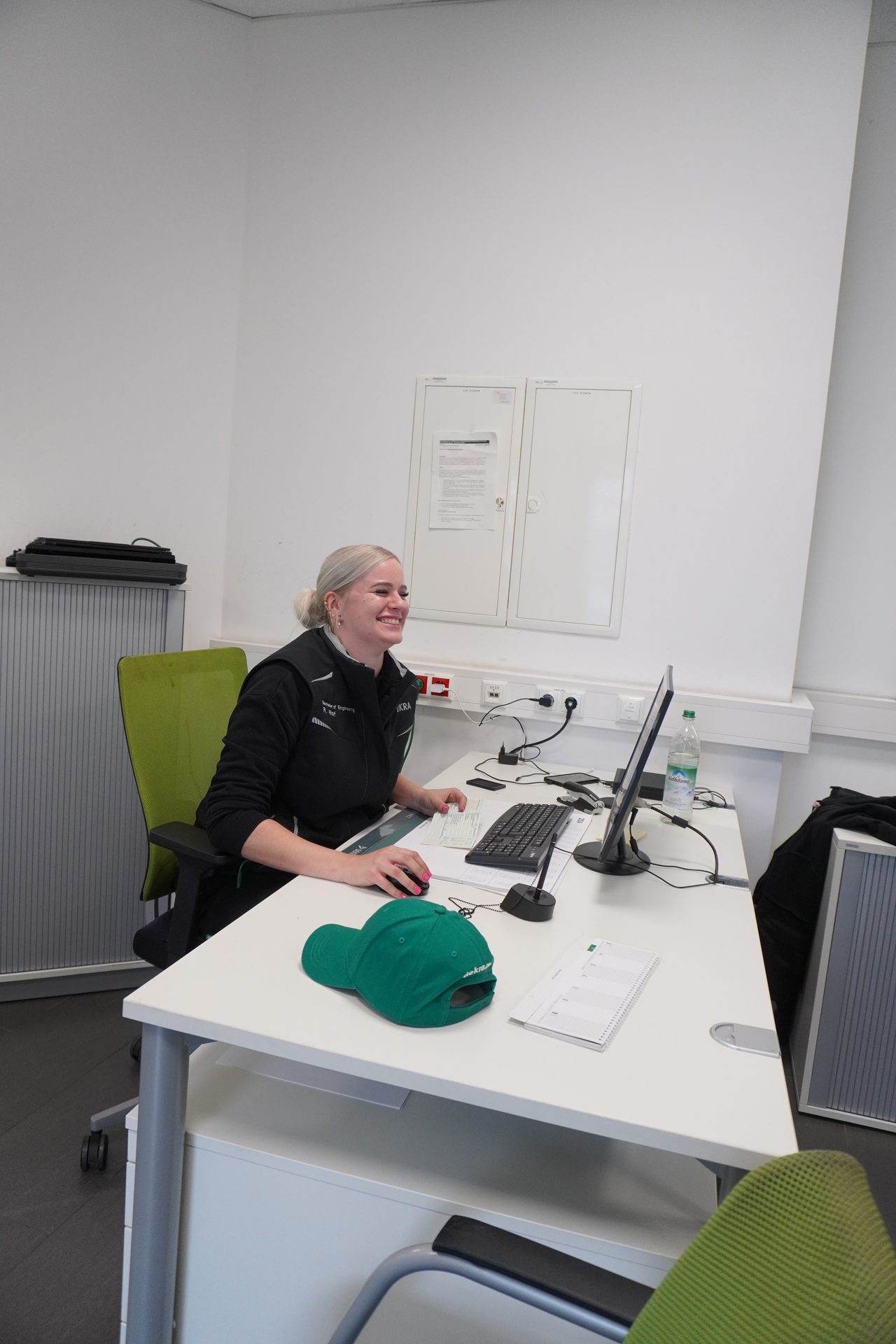A smiling woman with blonde hair in a ponytail works at a white desk with a computer monitor, keyboard, and mouse.