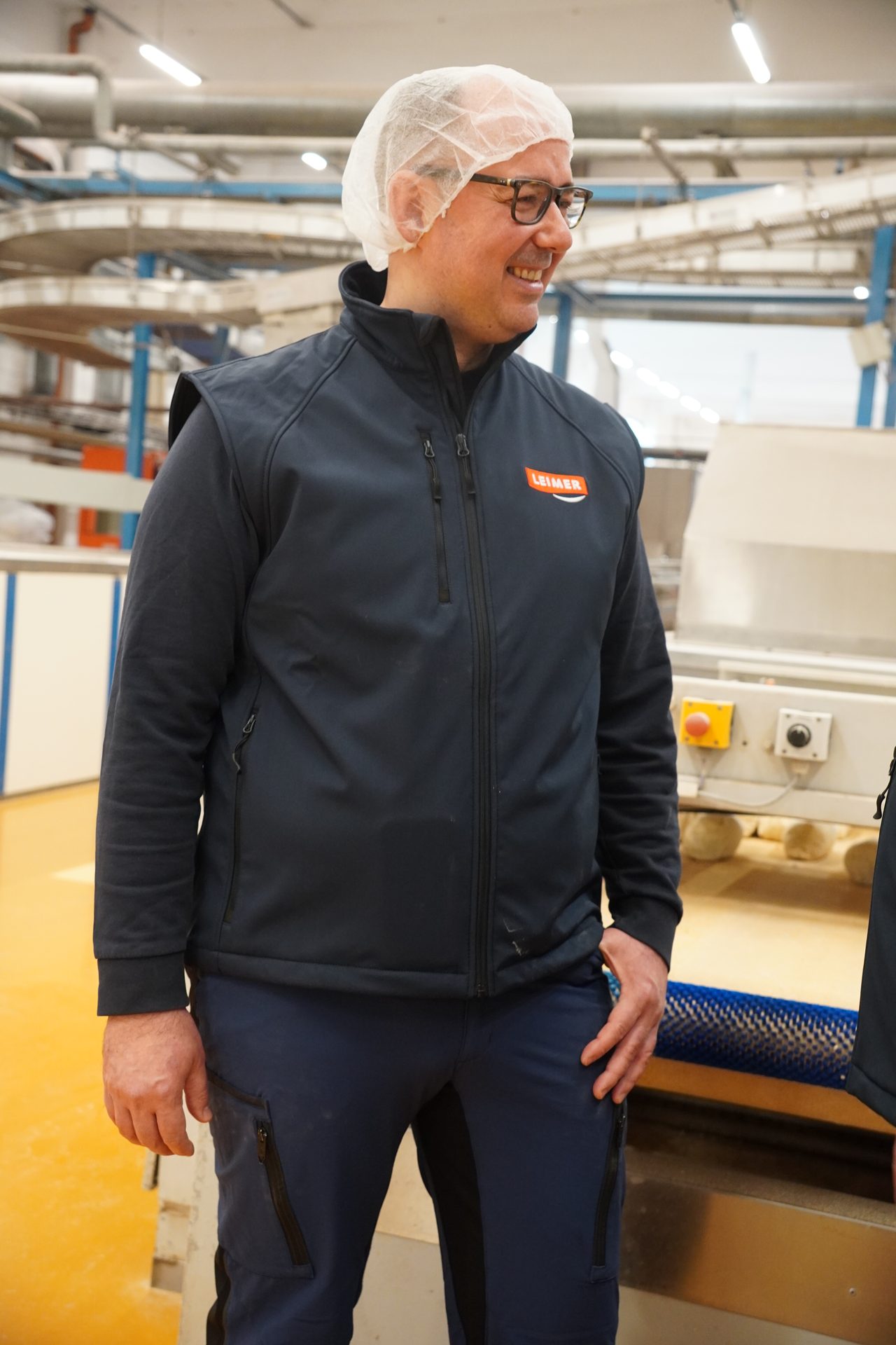 A smiling man wearing a hairnet and a blue vest with a "LEIMER" logo in a food production facility.