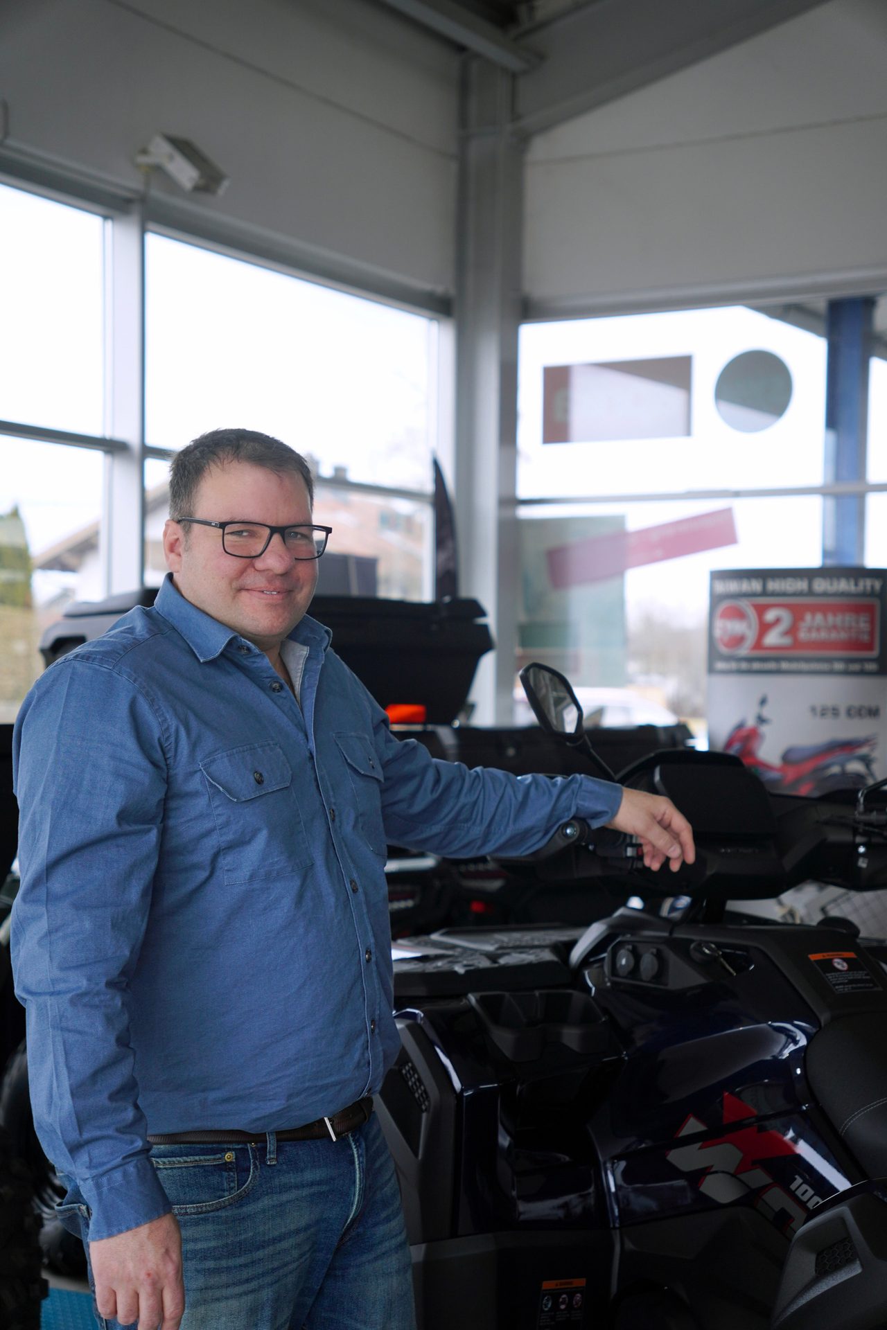 A smiling man in a blue denim shirt and jeans stands next to a dark quad bike in a vehicle showroom.