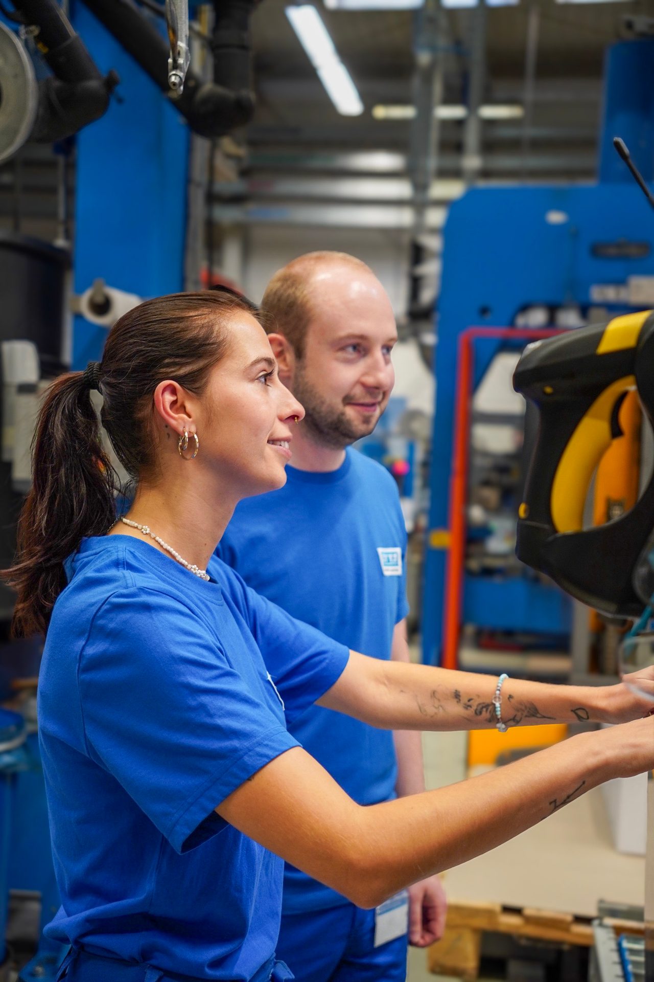 Two smiling factory workers in blue shirts operating machinery, focused on their task.