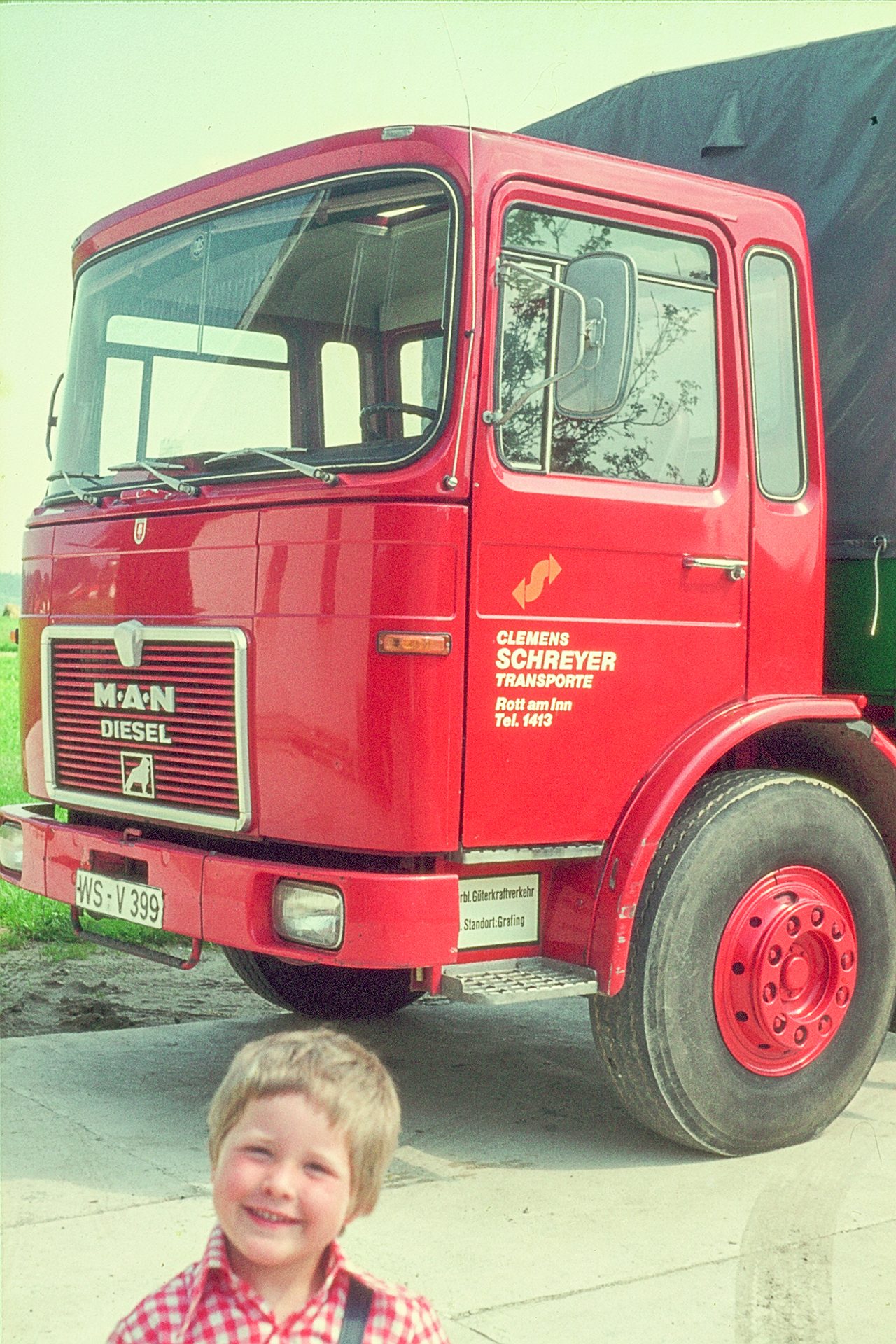 Young boy smiling in front of a red MAN Diesel truck belonging to Clemens Schreyer Transporte.