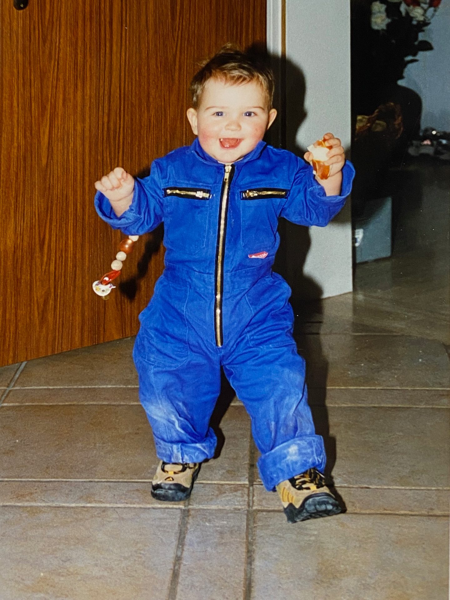 A happy toddler in a blue jumpsuit smiles at the camera, holding a piece of food and a beaded toy.