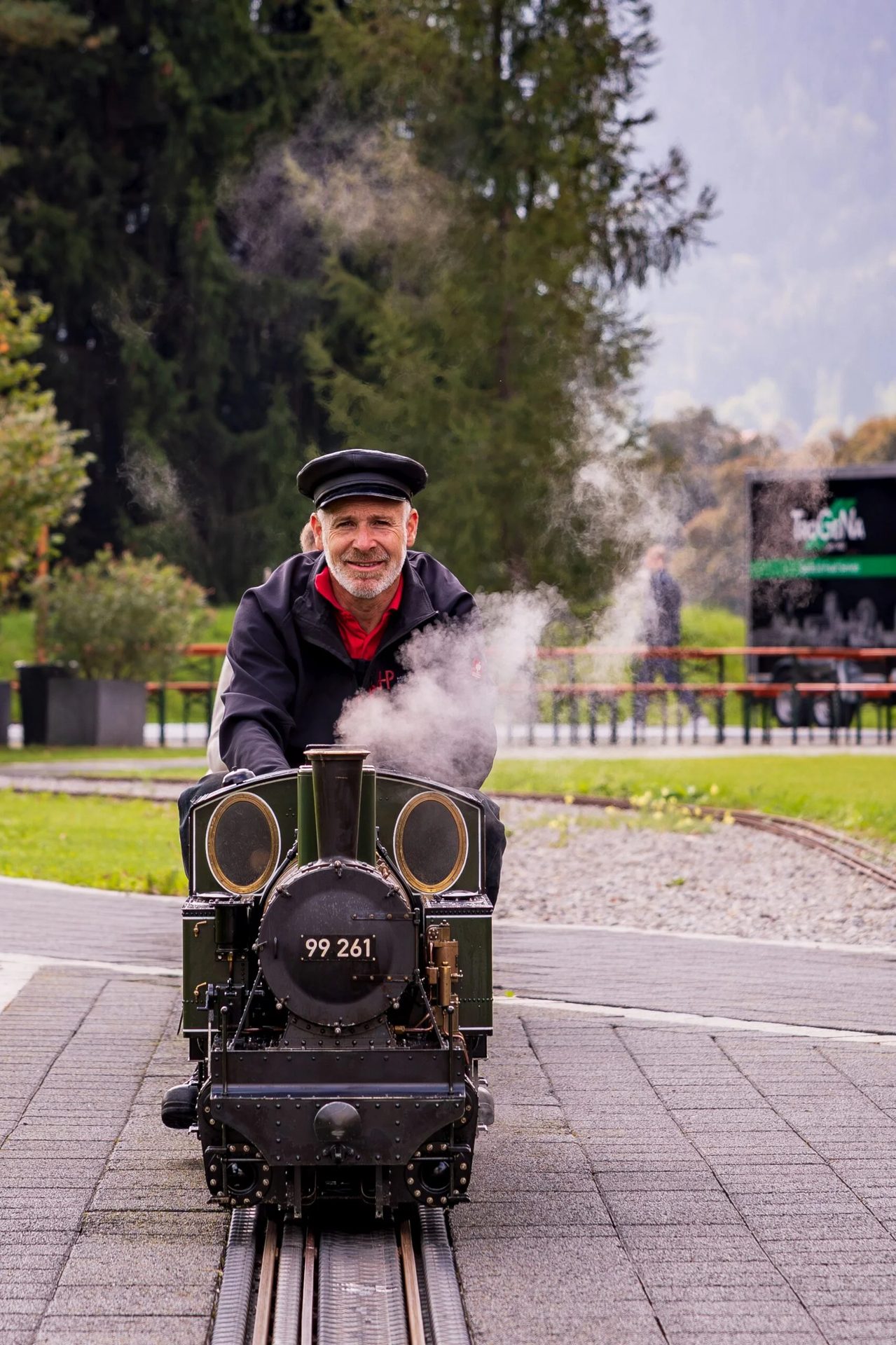 Smiling man in a cap drives a miniature steam train, puffing smoke, on outdoor tracks.