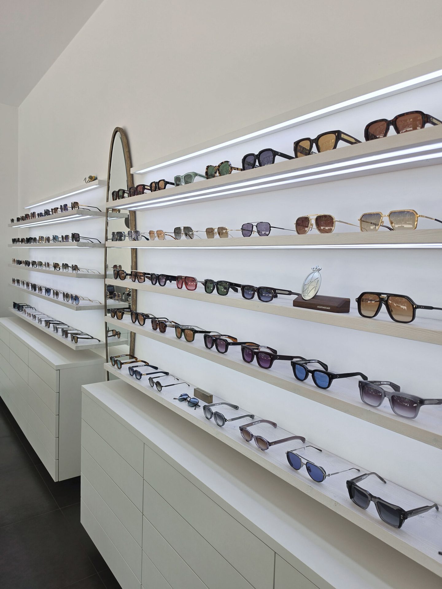 An optical store with multiple rows of sunglasses displayed on illuminated white shelves and cabinets.