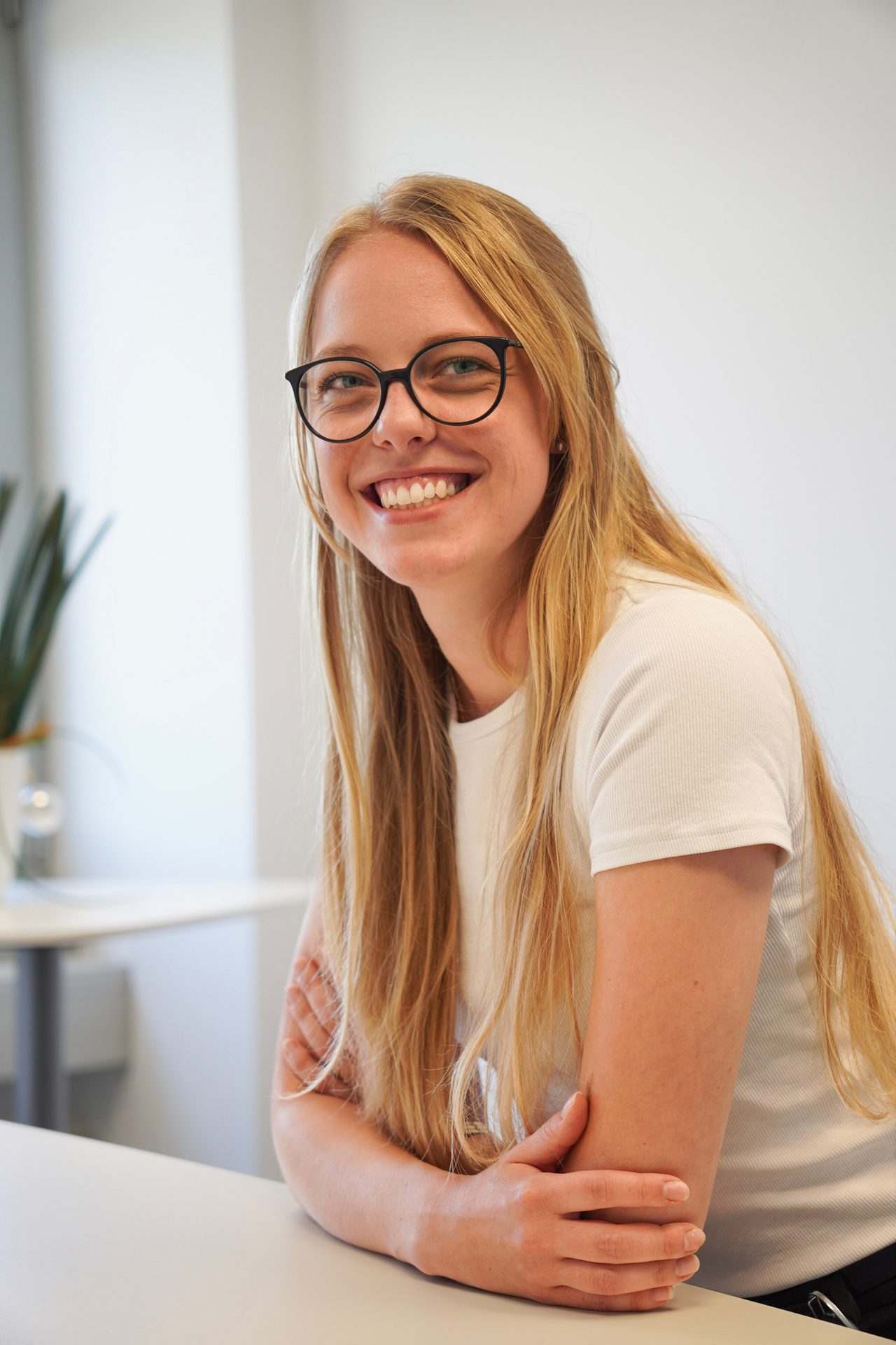 Smiling young woman with blonde hair and glasses leaning on a table.
