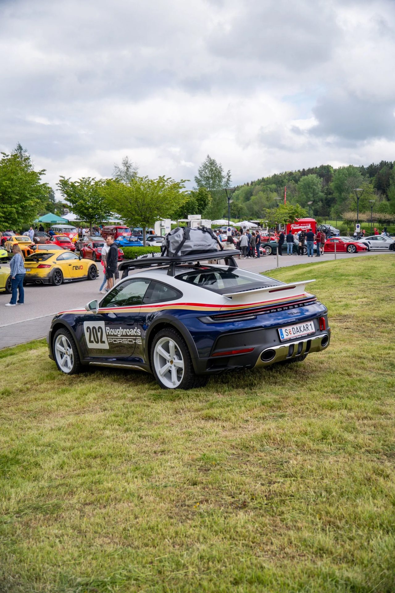 A blue and white Porsche 911 Dakar with a roof rack and '202 Roughroads' livery parked on grass at an outdoor car show.