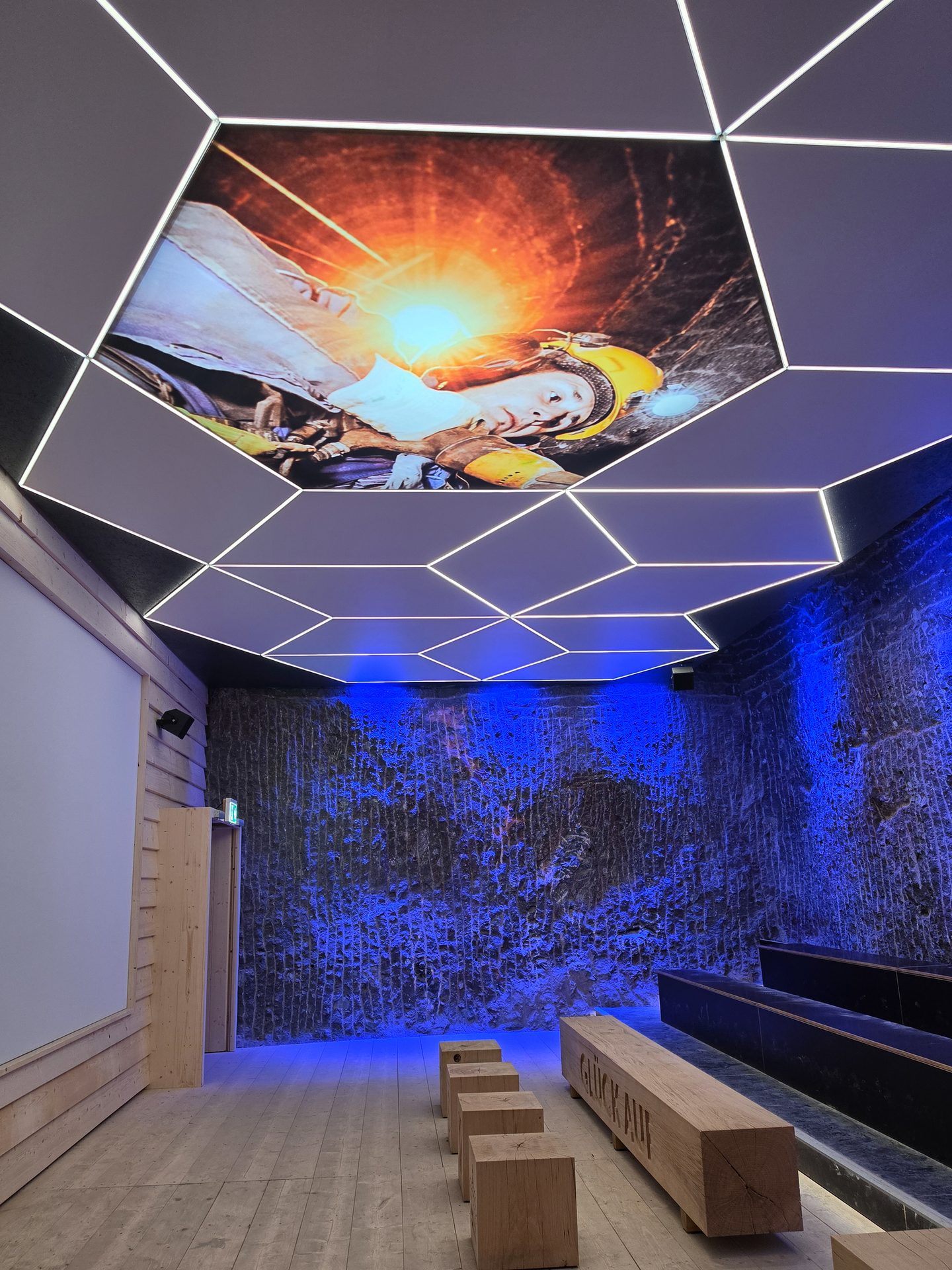 Indoor exhibit with hexagonal ceiling display of a welder, blue-lit textured wall, and wooden seating.