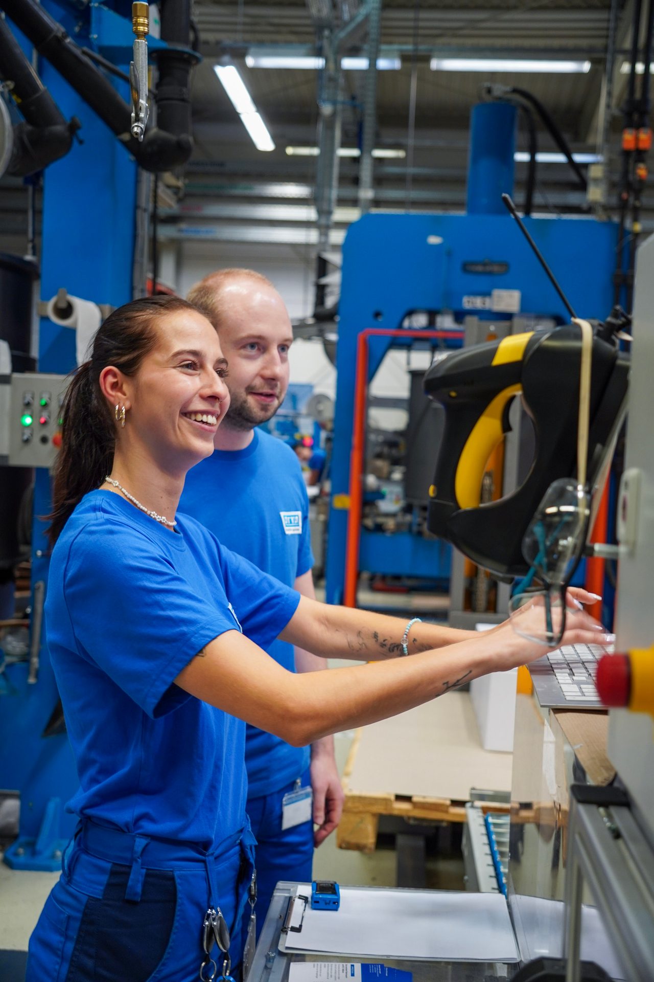 Two factory workers in blue shirts, a woman typing on a laptop and a man observing, smiling in a plant.