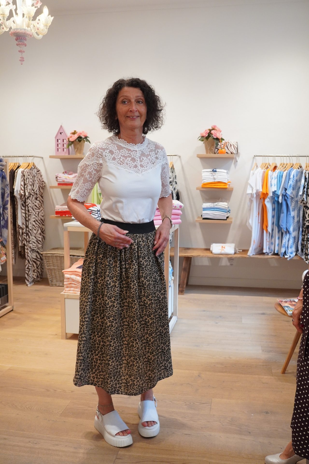 A woman in a white lace top and leopard print skirt poses in a clothing store, smiling slightly.