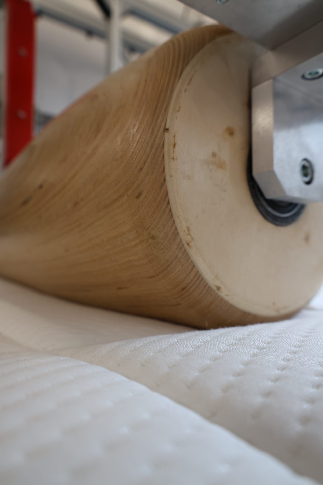 A textured roller, possibly wood, presses down on a white quilted fabric or mattress, with a metal bracket visible.