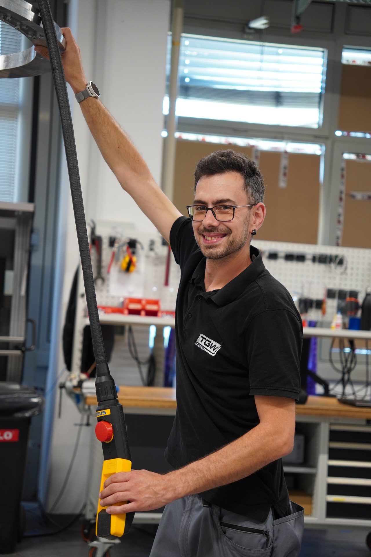 A smiling man in glasses and a black polo shirt, holding a black and yellow control device in a workshop.