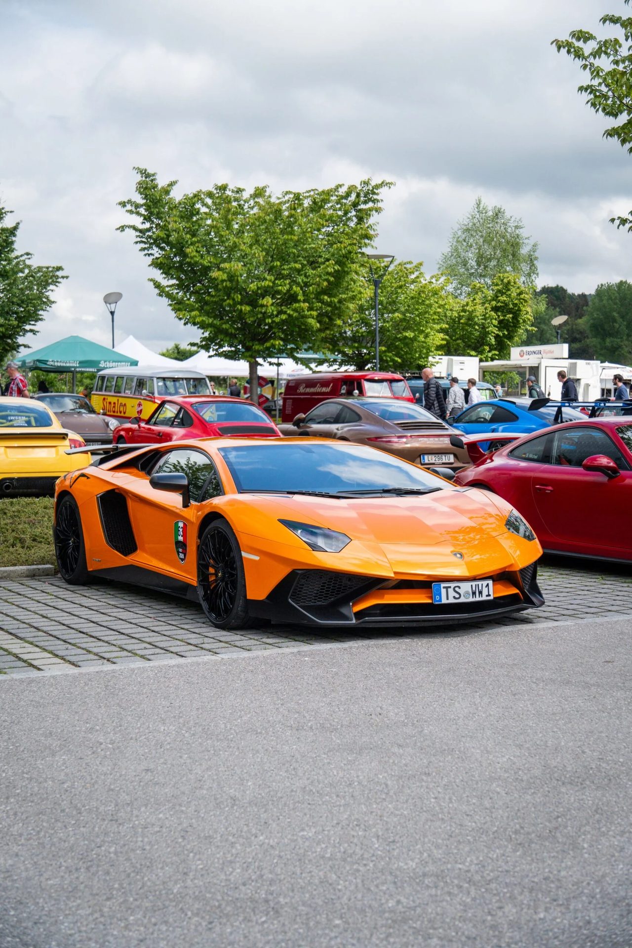A striking orange Lamborghini Aventador leads a display of luxury and classic cars at an outdoor show.