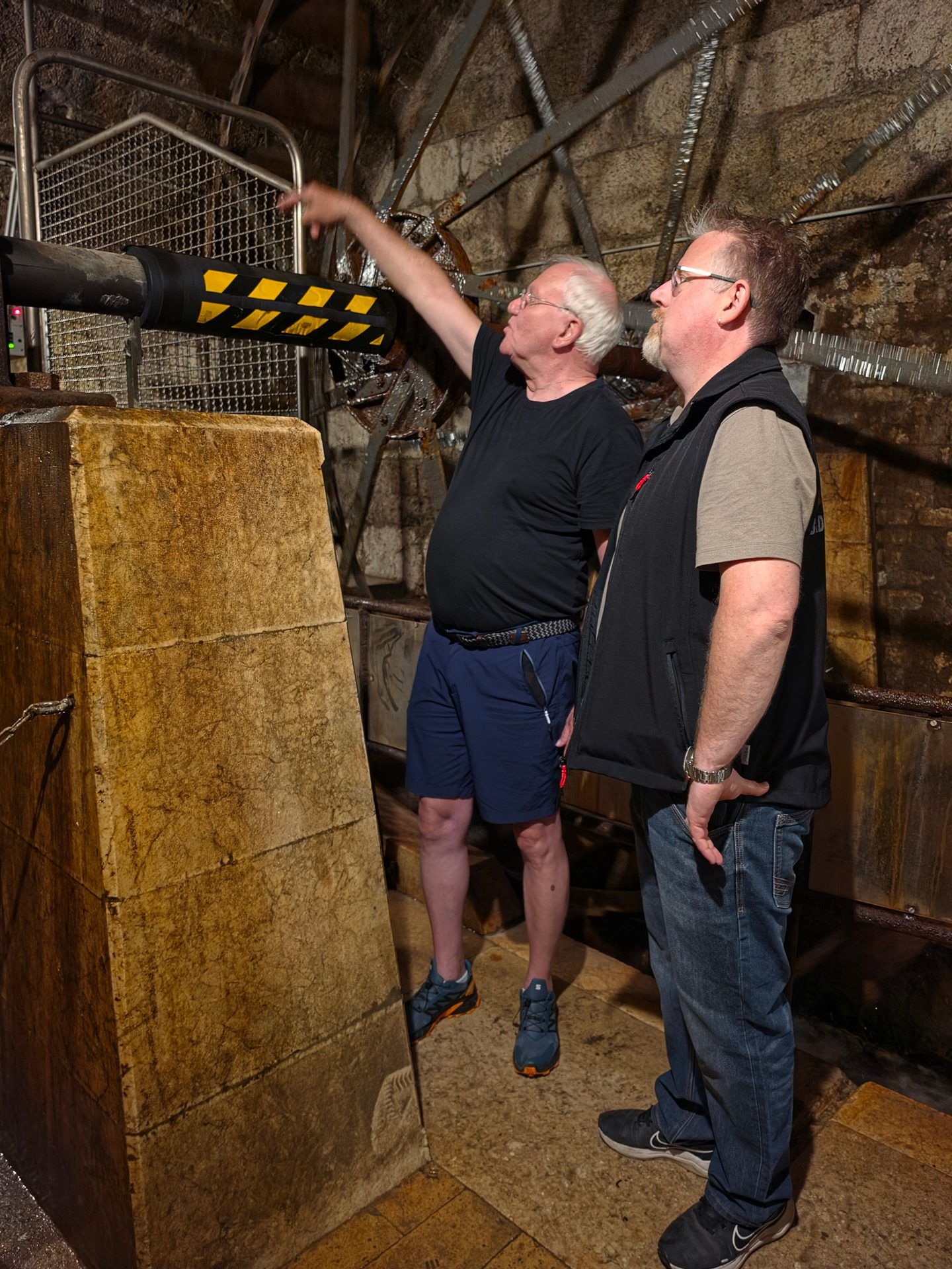 Two men observe large industrial machinery, with one pointing upwards in a rustic, dimly lit interior.