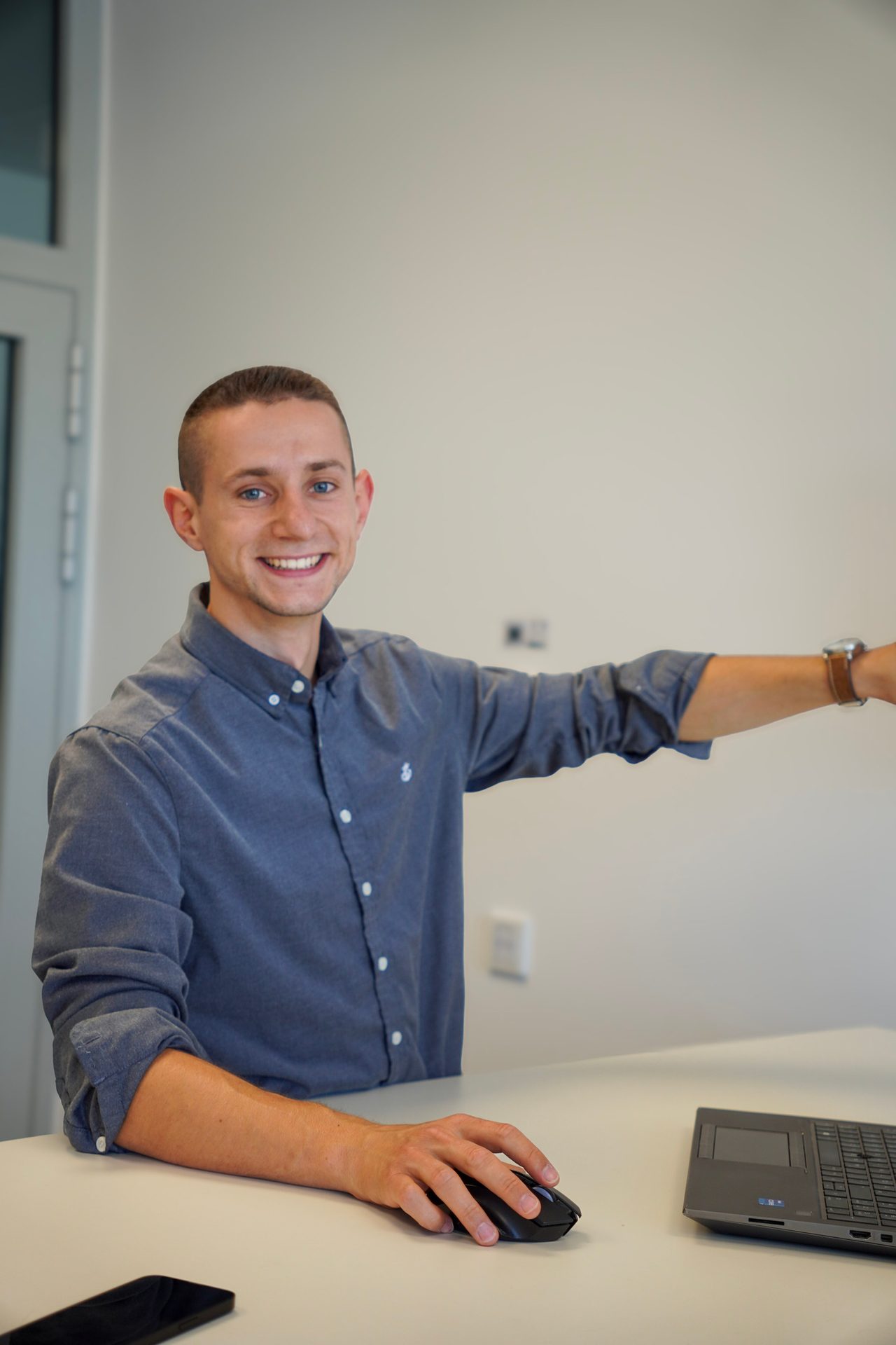 Young man smiling, pointing to the side, and using a computer mouse at a desk in an office.