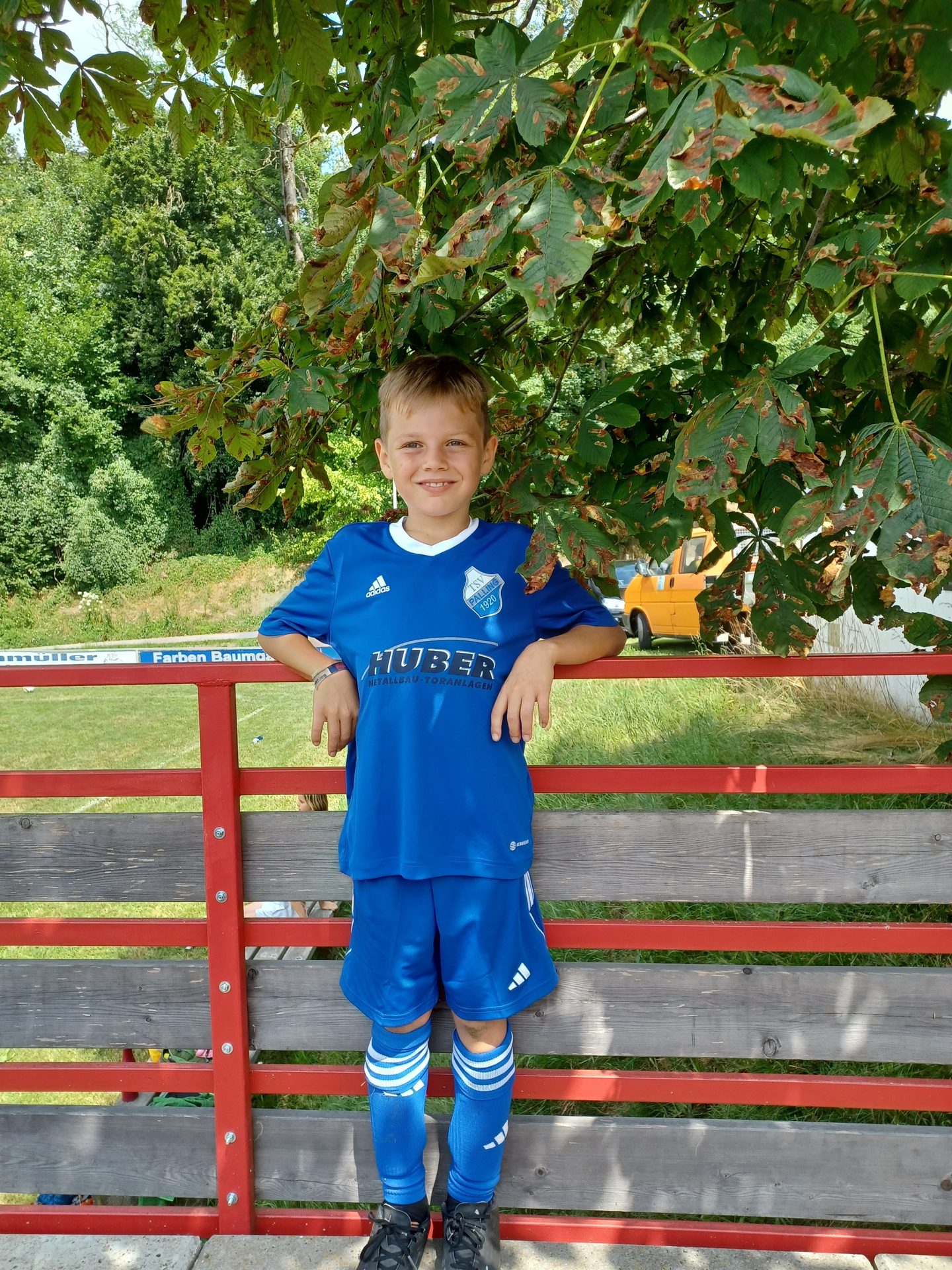 A young boy in a blue soccer uniform smiles happily while leaning on a red fence outdoors.