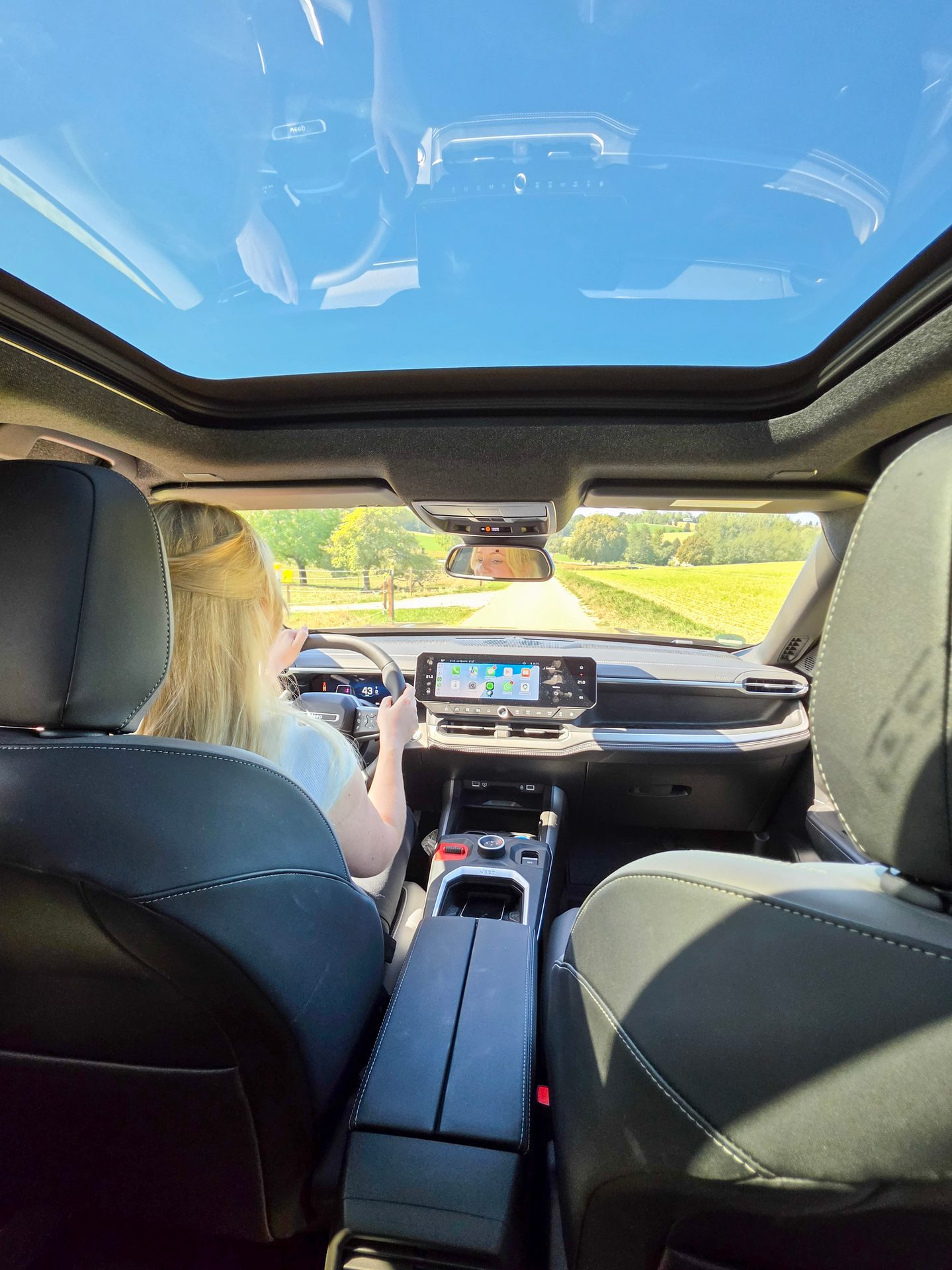 A person with blonde hair drives a modern car with a panoramic sunroof, looking ahead at a country road.