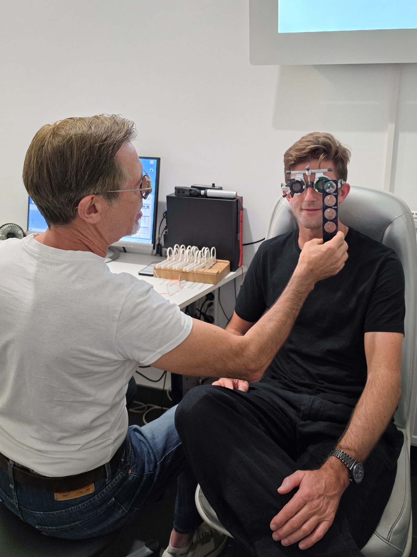 An optometrist performs an eye exam on a male patient using a trial frame and lenses.