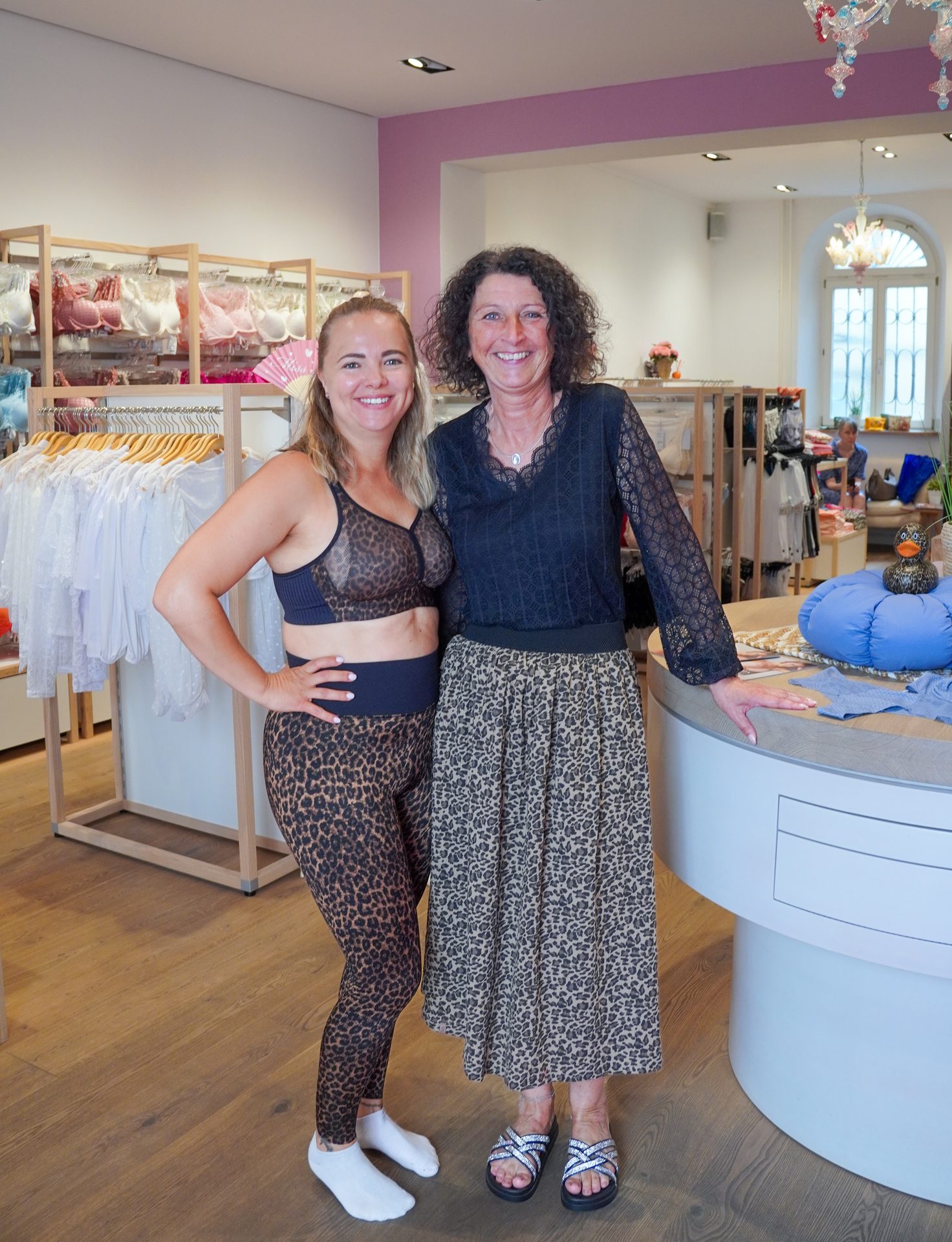 Two smiling women in leopard print clothing stand in a lingerie store, one in activewear, the other in a skirt.