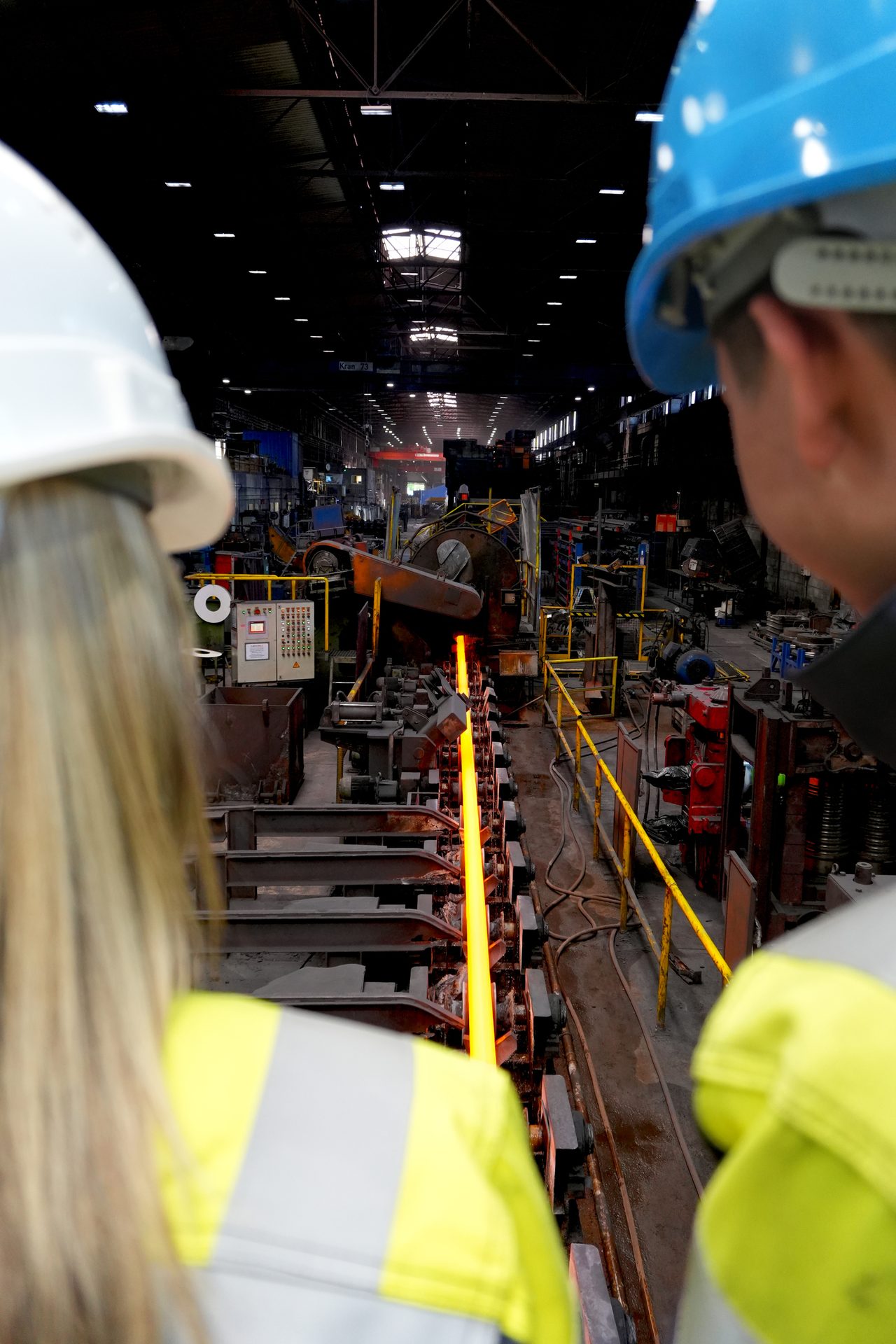 Two workers in hard hats observe glowing hot metal being processed in a steel rolling mill.