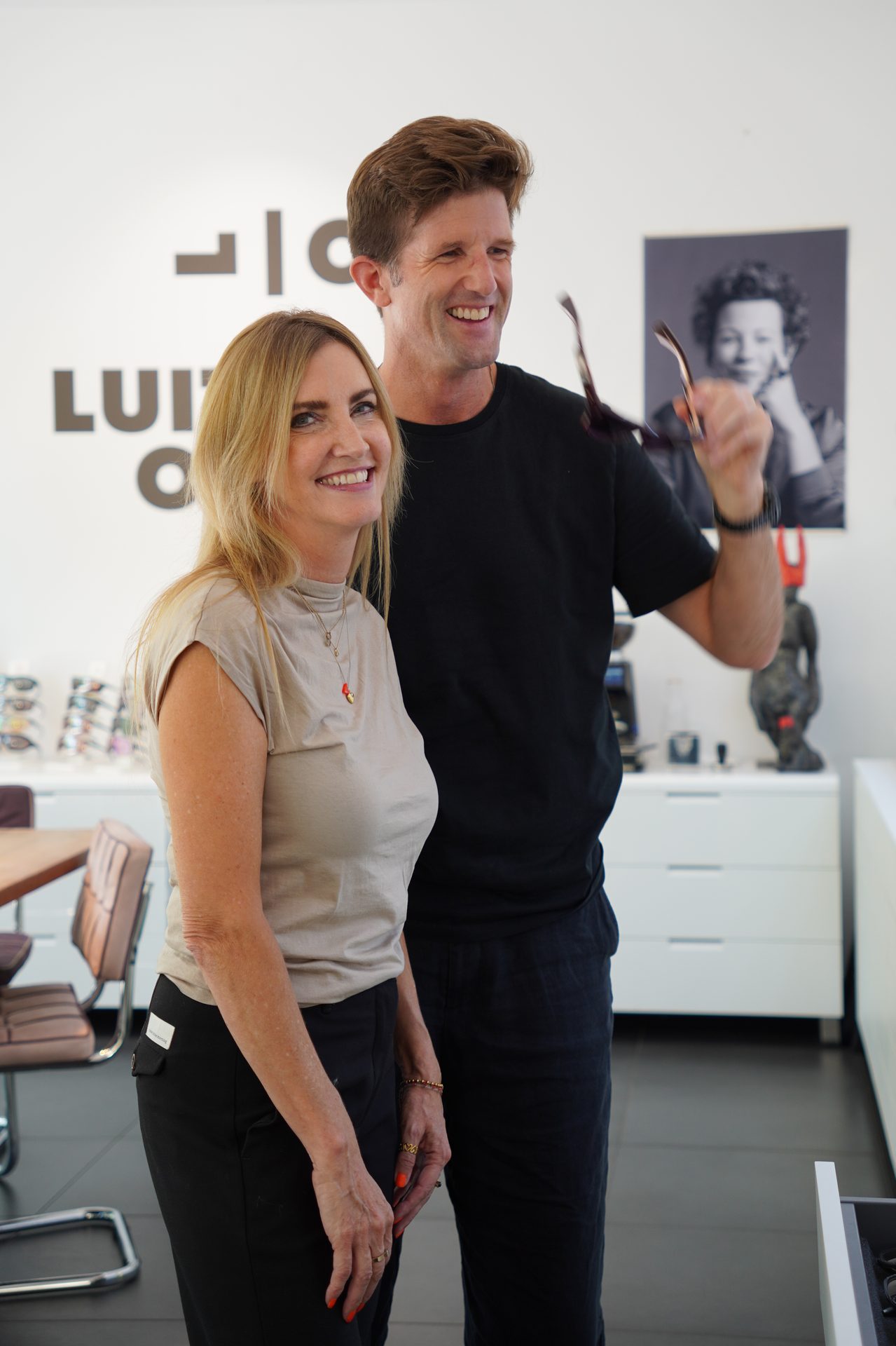 A man holds glasses, smiling with a woman in an optician's store, both happy and engaged.