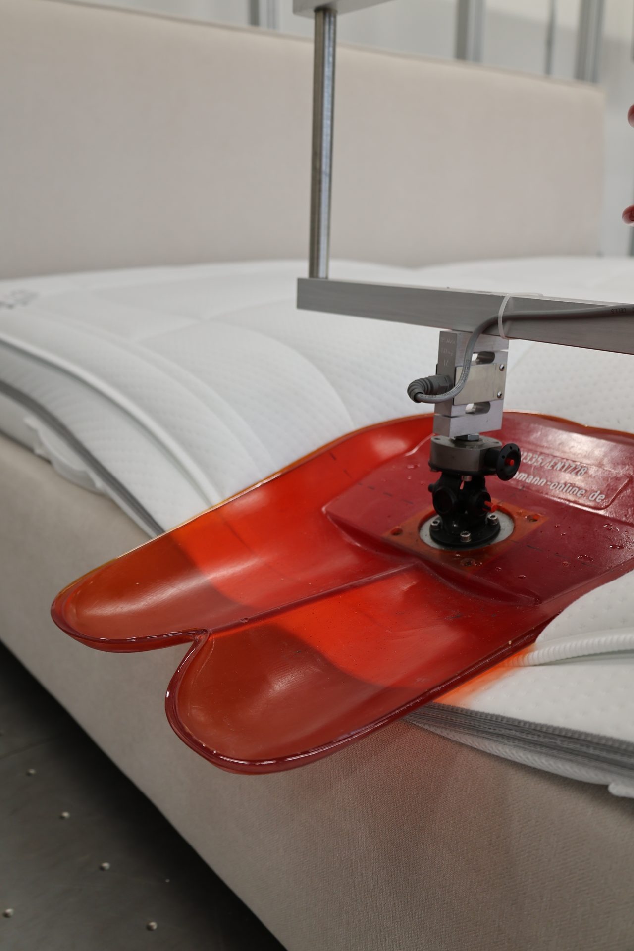 Red, foot-shaped device pressing on a white mattress for durability testing.
