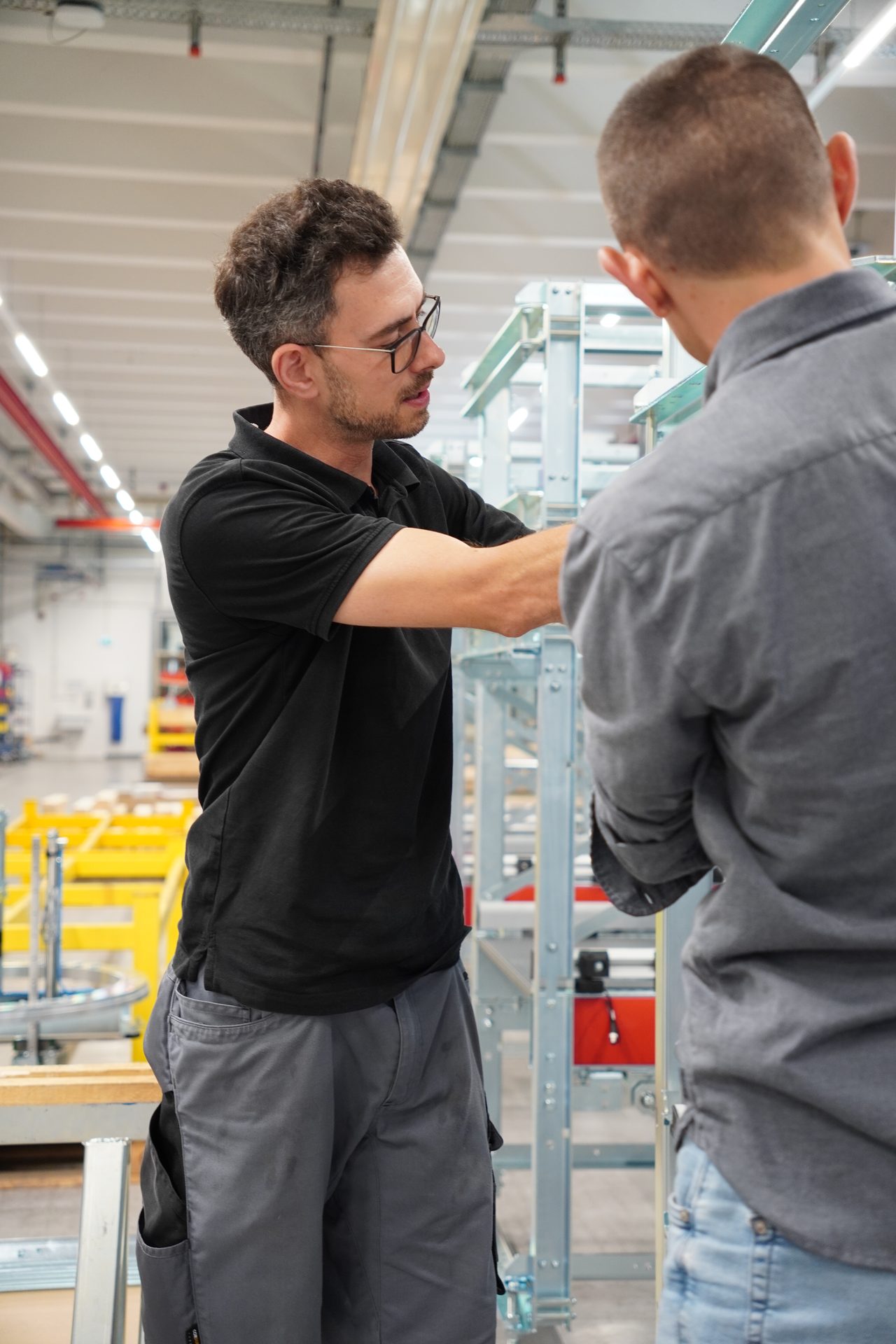 Two men work on industrial machinery in a factory, one explains while the other listens.