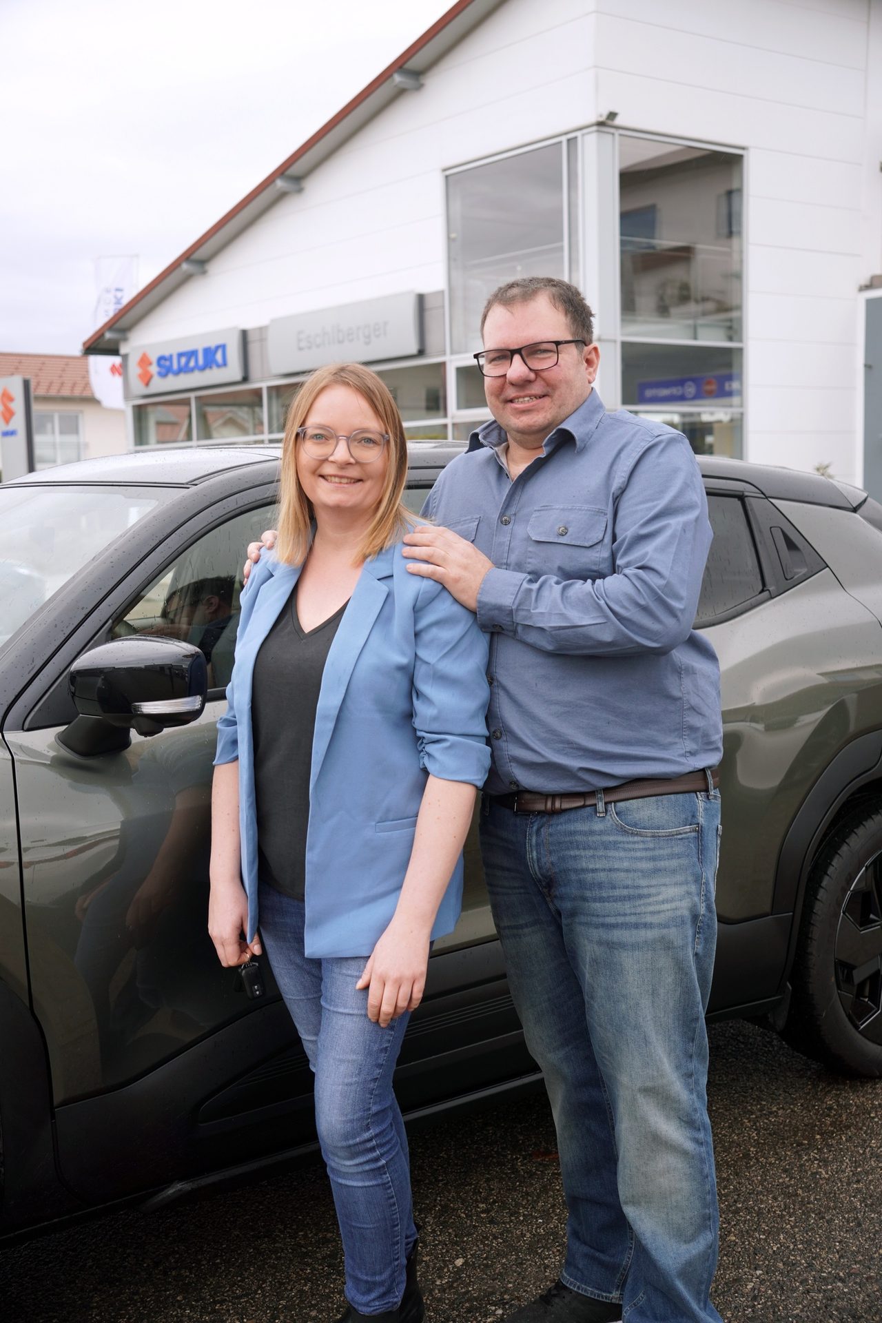A happy couple stands next to a new car in front of a Suzuki dealership building.