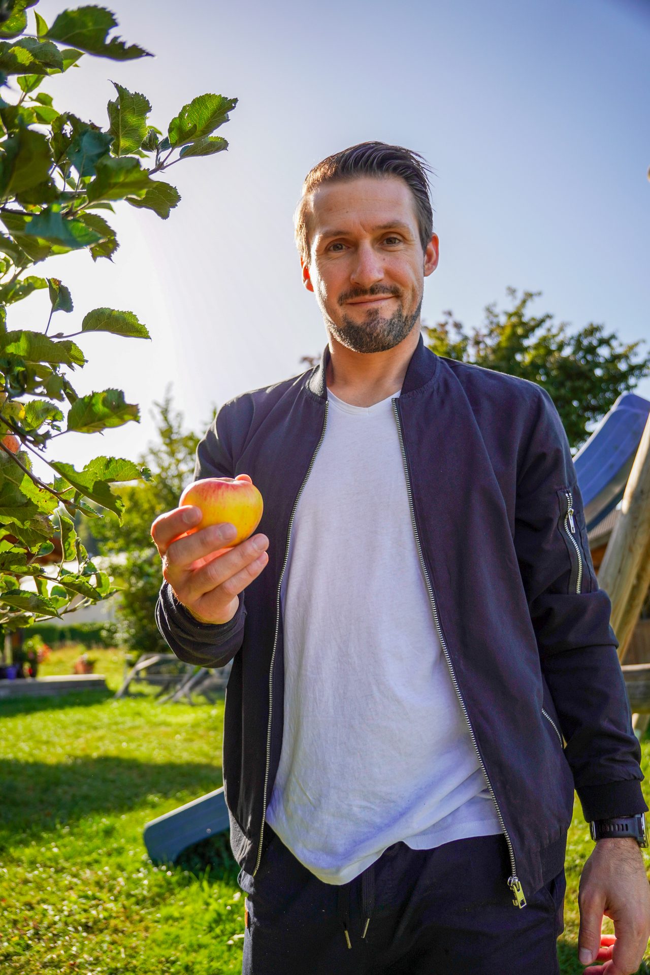 A man holding a fresh apple in a sunny outdoor setting with green foliage and clear sky.