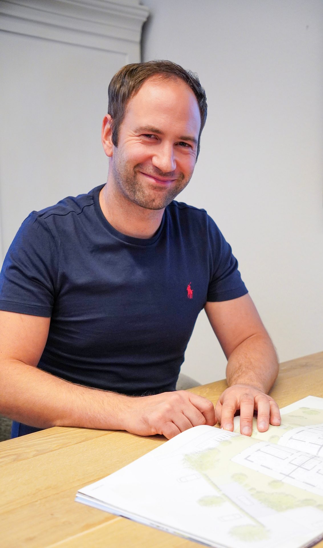 A man in a navy blue t-shirt smiles at the camera while sitting at a table with papers.