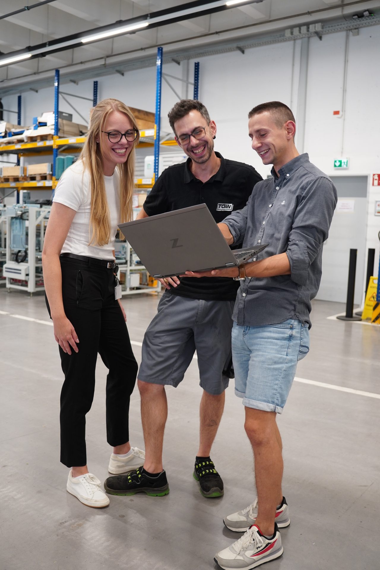 Three smiling colleagues collaborating over a laptop in a bright industrial or warehouse setting.