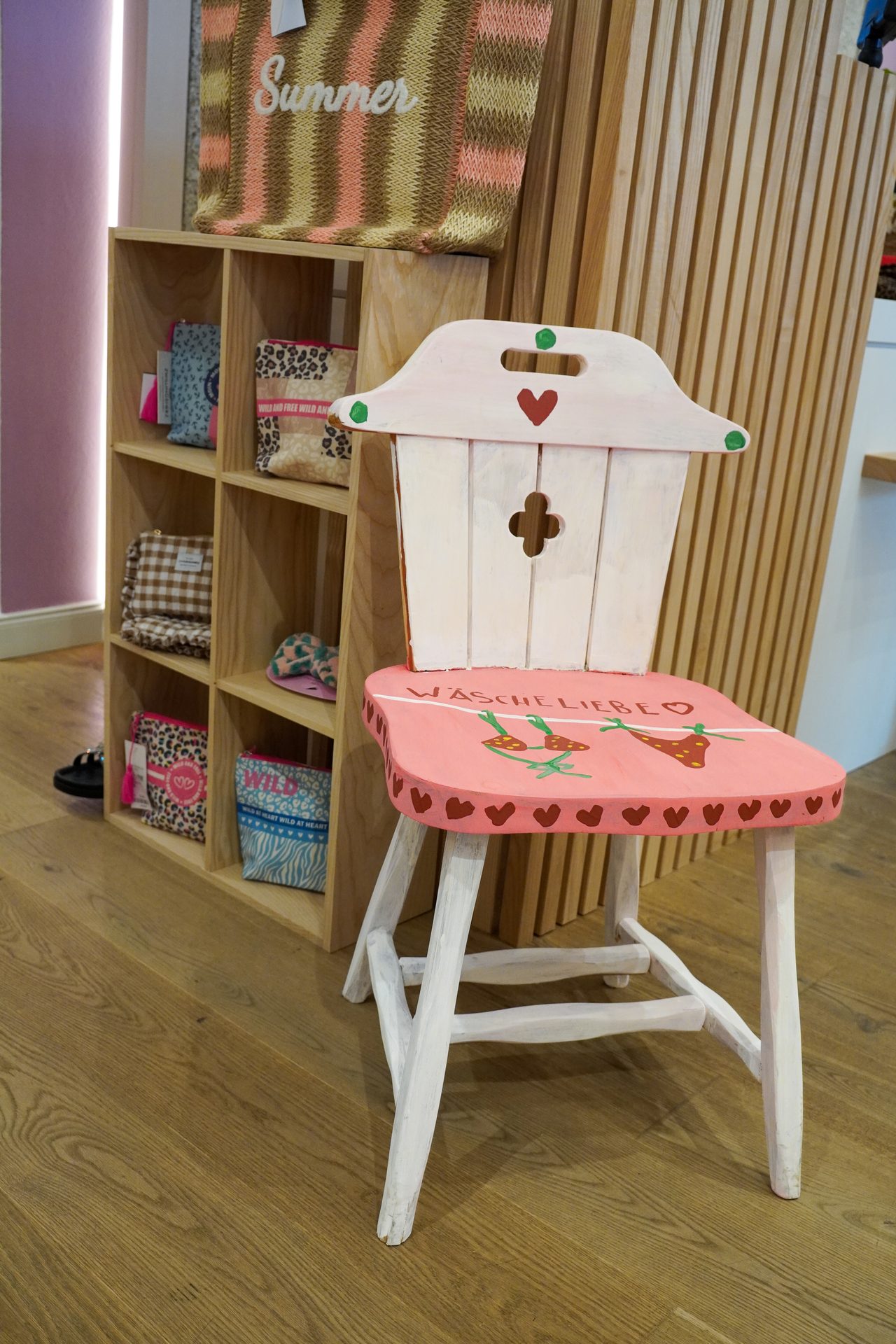 Pink decorated chair with "Wäscheliebe" text, next to a wooden shelf with patterned bags and pouches.