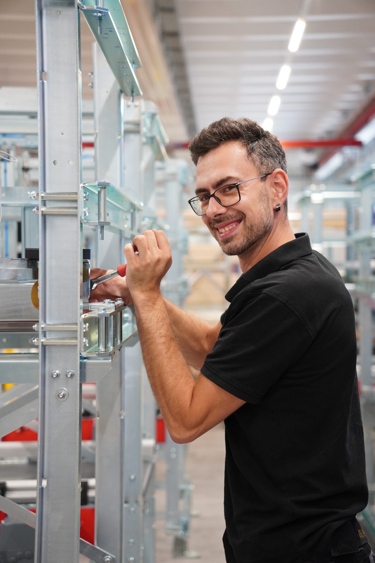 A smiling male worker in black polo shirt and glasses adjusts machinery with a tool in a brightly lit factory.