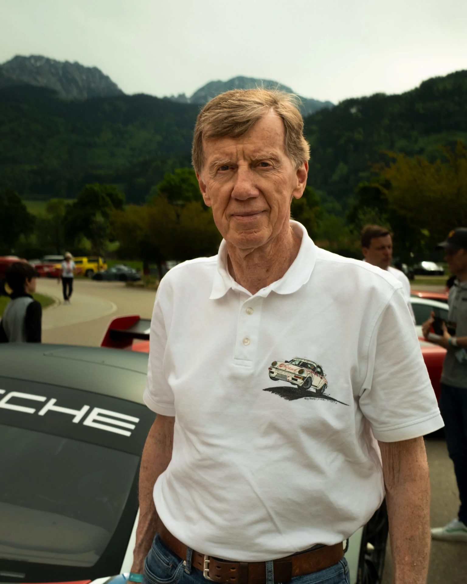An older man in a white polo shirt with a Porsche graphic stands outdoors, mountains visible.