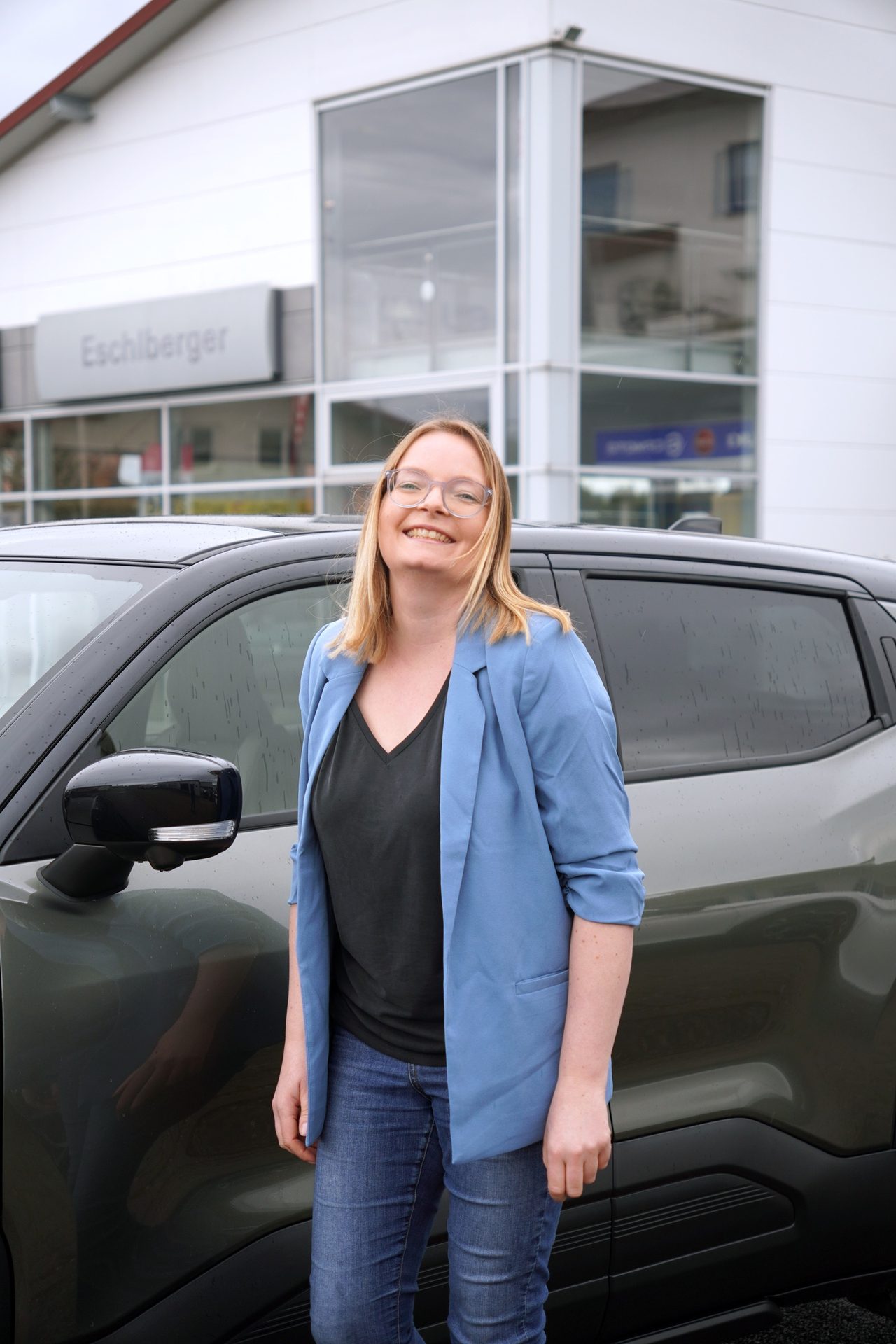 Smiling woman in glasses and blue blazer next to a dark car outside a dealership.