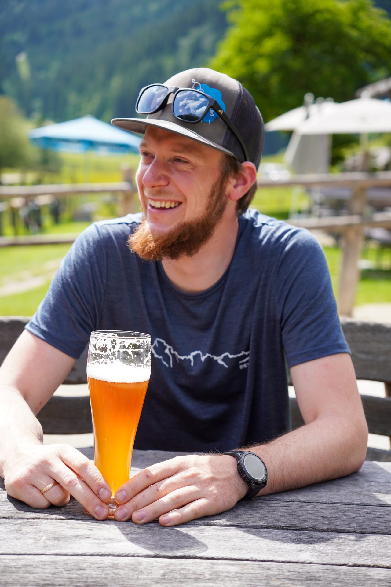 A smiling man with a beard and cap holds a glass of beer at an outdoor table on a sunny day.