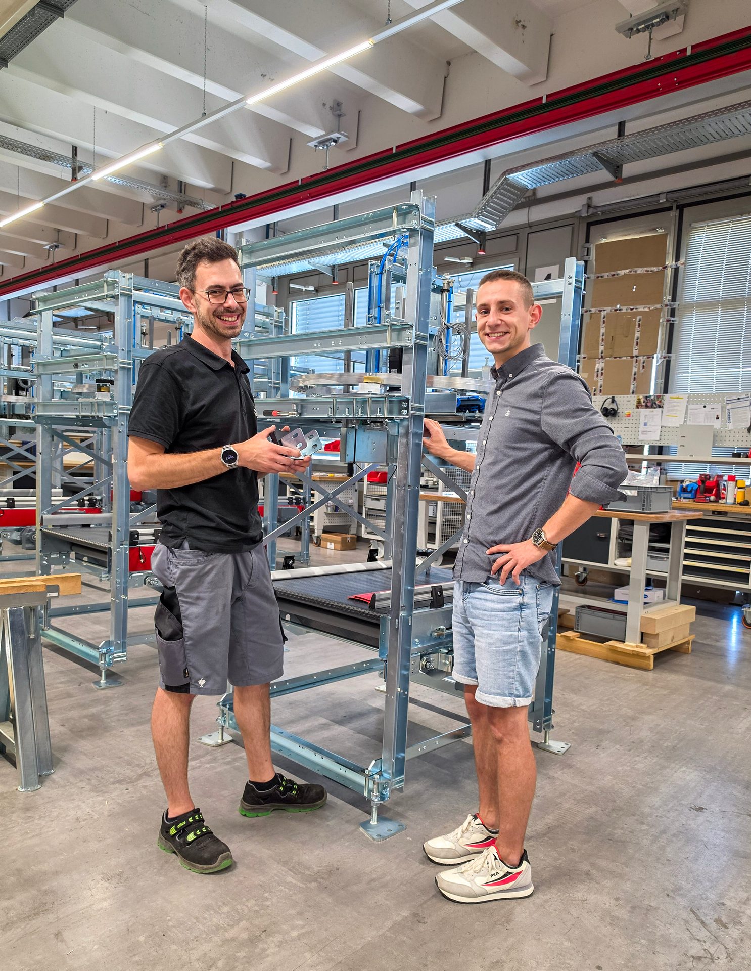 Two smiling men stand in a factory workshop, surrounded by metal structures and machinery.