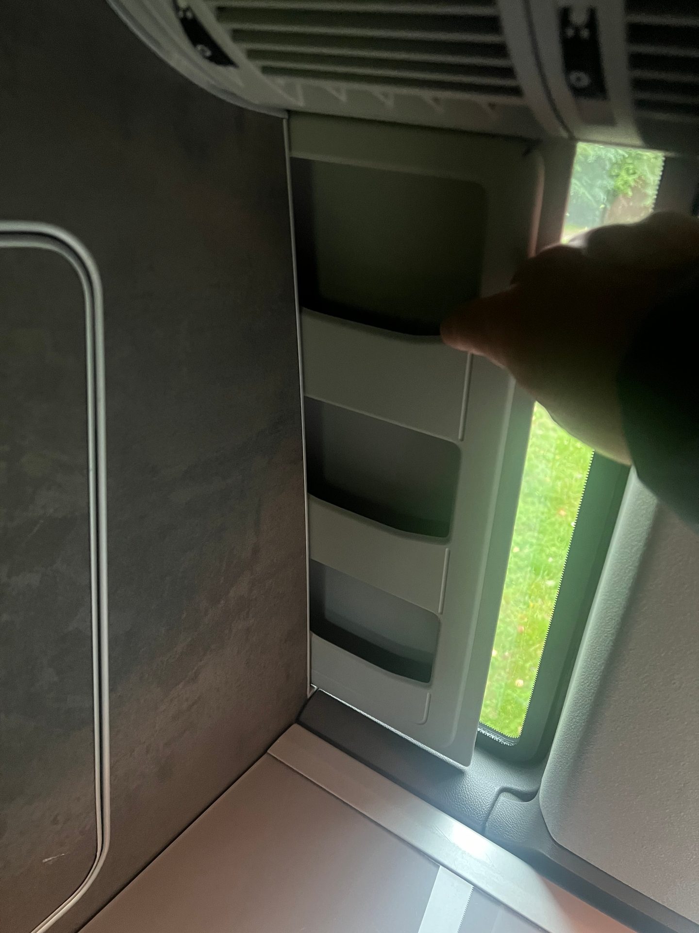Hand reaching into a vehicle's tiered storage by a window showing green outside.