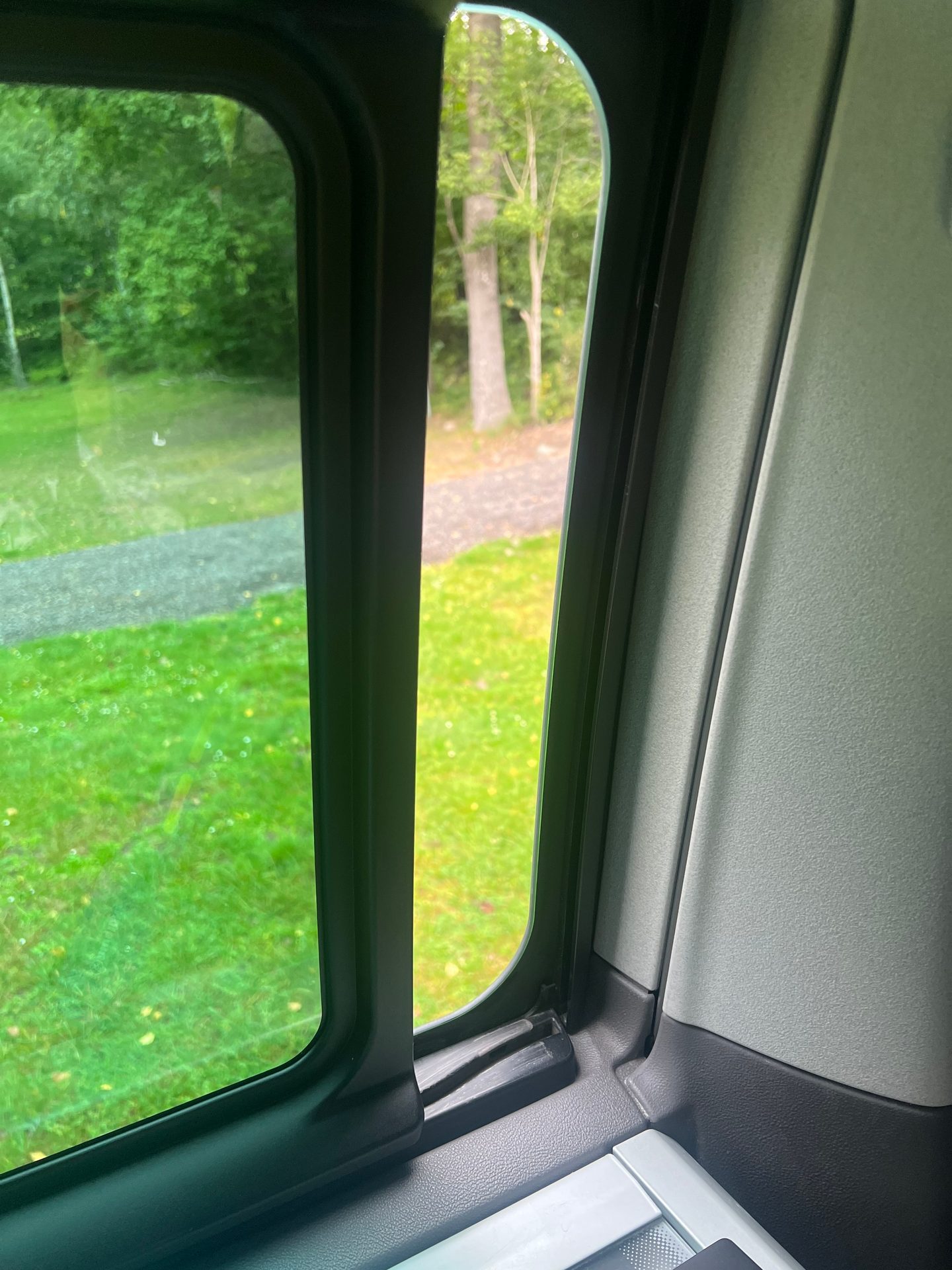View from inside a vehicle, looking through a side window at a green outdoor scene with trees and a path.