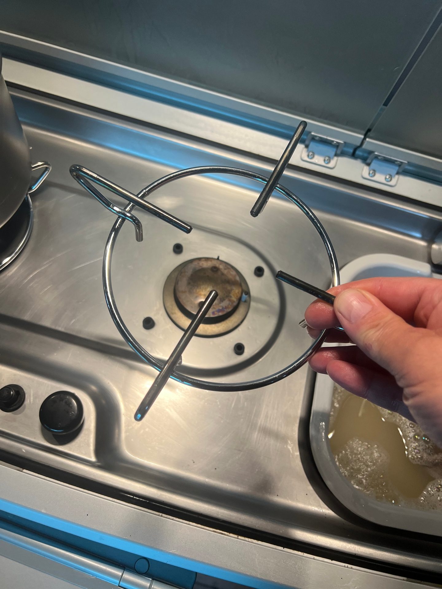 Person holding a metal trivet over a gas stove burner and sink with soapy water.