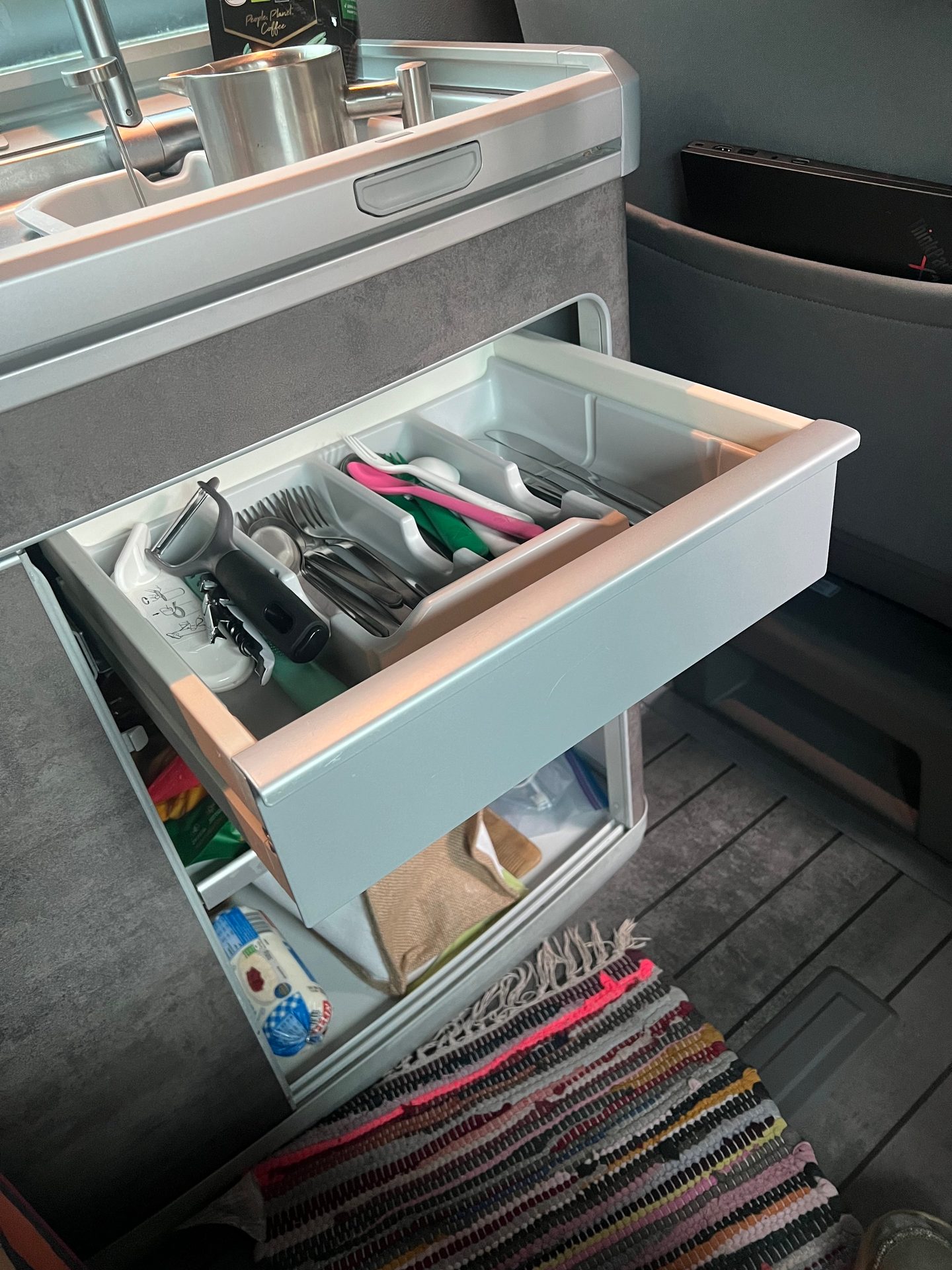 Compact kitchenette with open drawers, cutlery, sink, coffee, and a colorful rug on the floor.